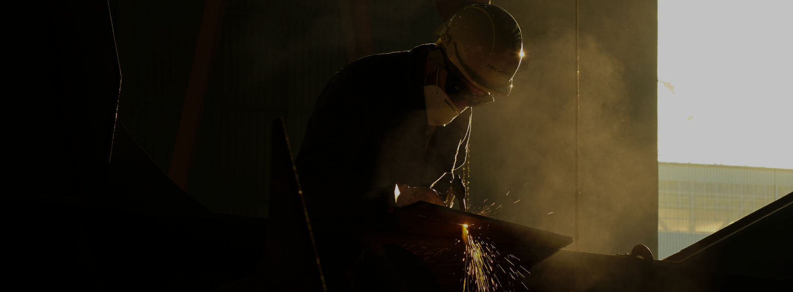 Fincantieri - People: Worker wearing a helmet uses a welding tool on a metal plate in backlight.