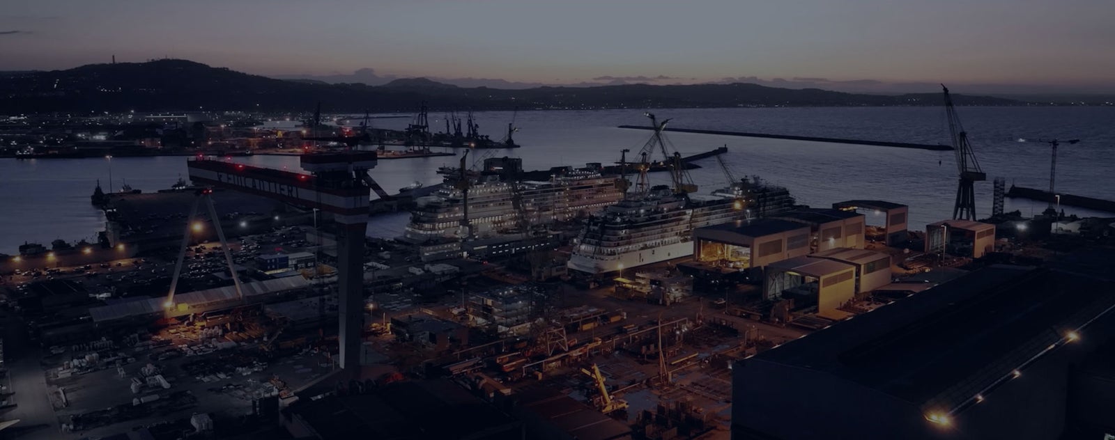 Fincantieri | About Us: Background of a port at sunset. The image is toned in dark shades close to black. It serves as the background for a banner.