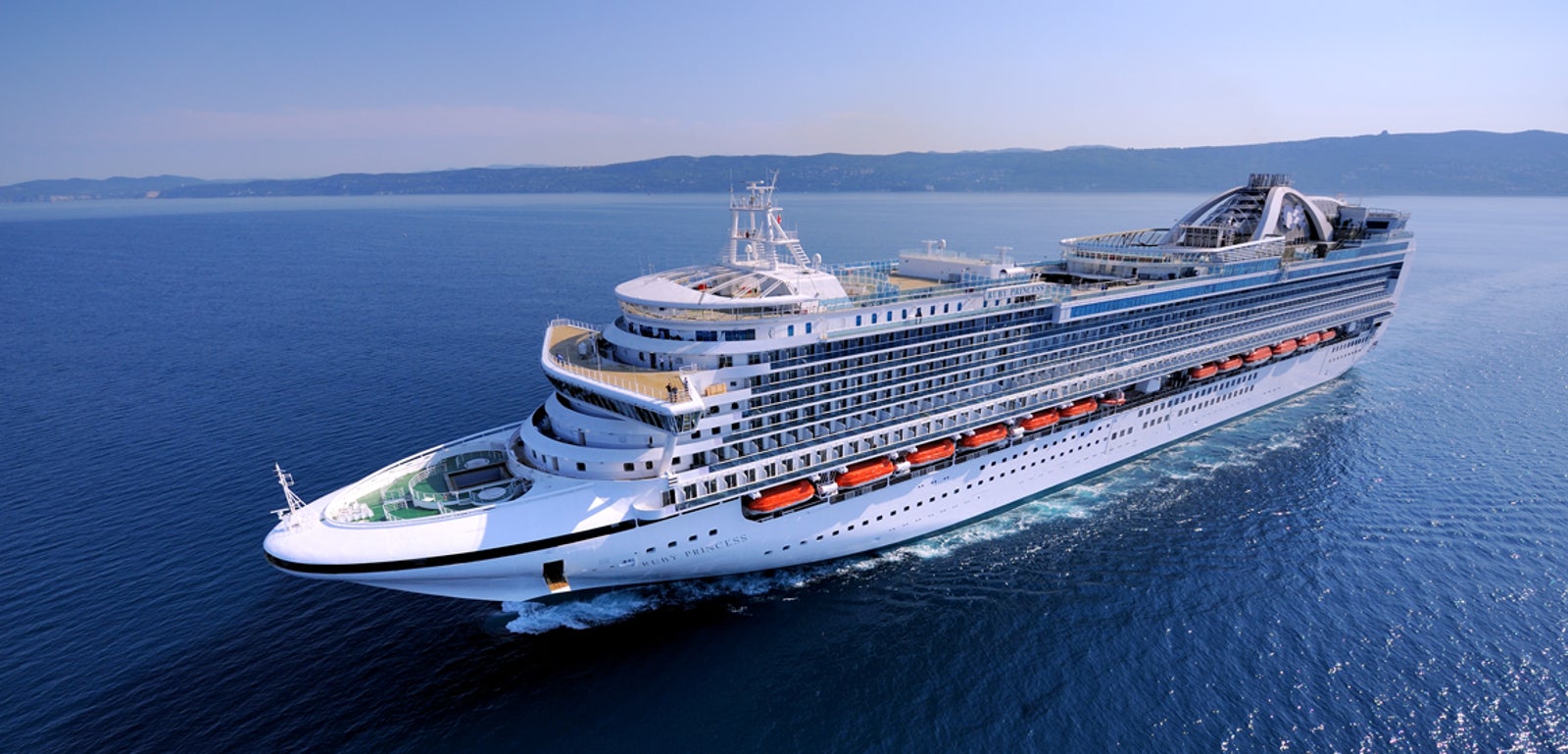 Fincantieri | Gruppo - Business - Prodotti - Navi da Crociera - Ruby Princess: Grande nave da crociera moderna operativa che scivola su acque calme e aperte sotto cielo azzurro limpido, più ponti, scialuppe di salvataggio e ampie finestre, maestosità e capacità di viaggio di piacere evidenziate.