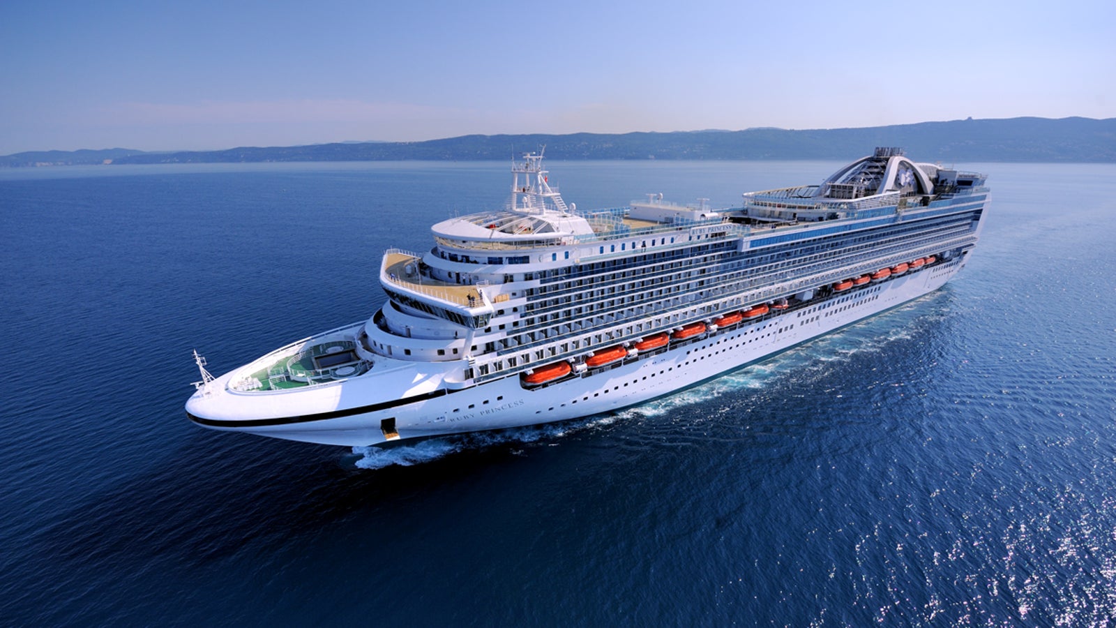 Fincantieri | Gruppo - Business - Prodotti - Navi da Crociera - Ruby Princess: Grande nave da crociera moderna operativa che scivola su acque calme e aperte sotto cielo azzurro limpido, più ponti, scialuppe di salvataggio e ampie finestre, maestosità e capacità di viaggio di piacere evidenziate.
