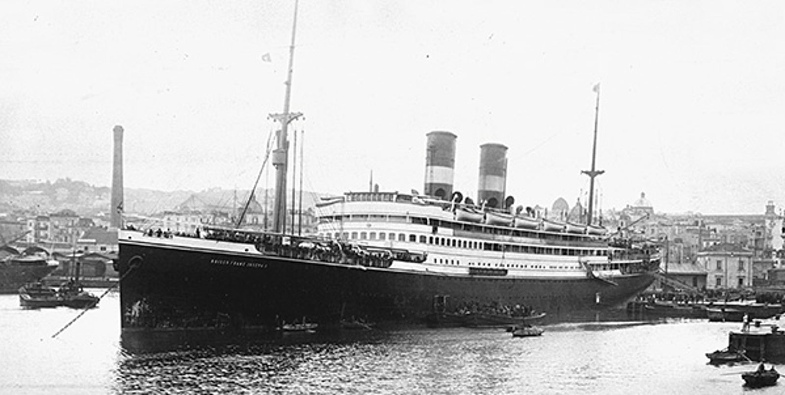 Fincantieri | About Us - Historic Ships: Large steamship with two funnels docked in a crowded port, surrounded by smaller boats, black and white photo with urban buildings in the background, the name 'Santa Rosa' clearly visible on the hull.