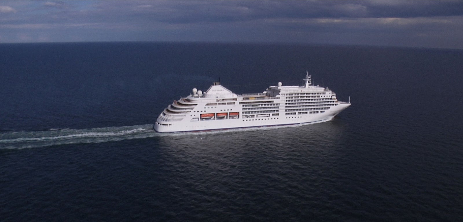 Fincantieri | Group - Business - Products - Cruise Ships - Silver Spirit: Large modern operational white cruise ship with multiple decks and visible lifeboats, sailing on calm waters under partly cloudy sky, visible wake accentuates movement, scene creates atmosphere of luxury, relaxation, and maritime adventure.
