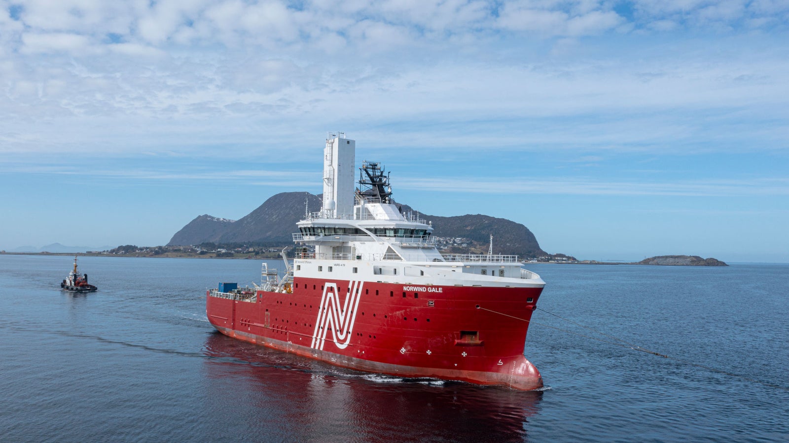 Prodotto - Offshore Support Vessel Norwind Gale: Vista laterale della nave operativa in navigazione, scafo rosso con grande logo N bianco, sfondo costiero montuoso e cielo sereno, rimorchiatore vicino.