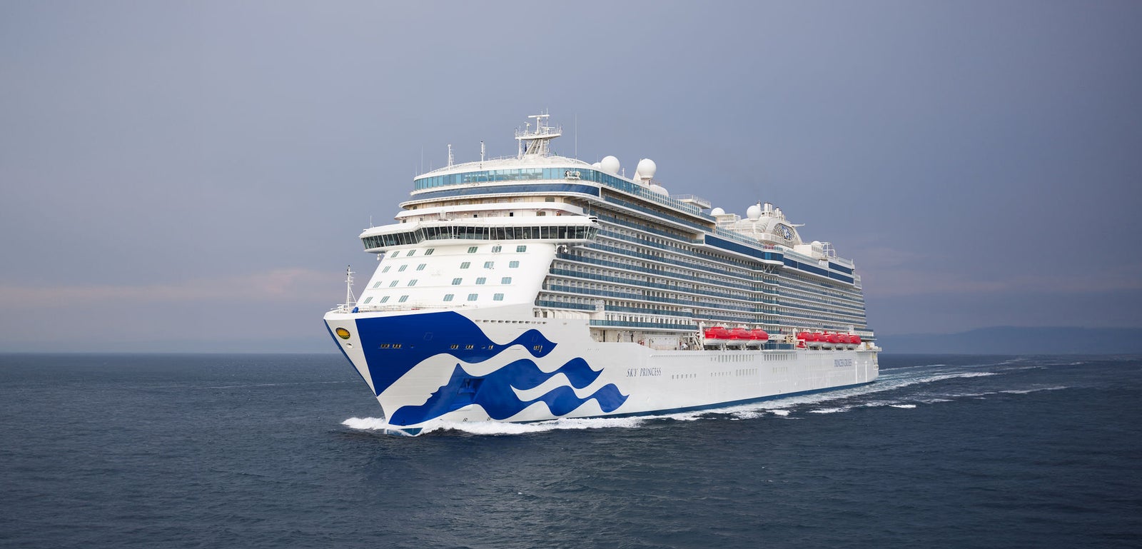 Fincantieri | Gruppo - Business - Prodotti - Navi da Crociera - Sky Princess: Grande nave da crociera di lusso operativa etichettata 'Sky Princess', naviga su acque calme dell'oceano sotto cielo nuvoloso, design elegante con grafica a onda blu sullo scafo, più ponti visibili, scialuppe di salvataggio rosse lungo i lati.