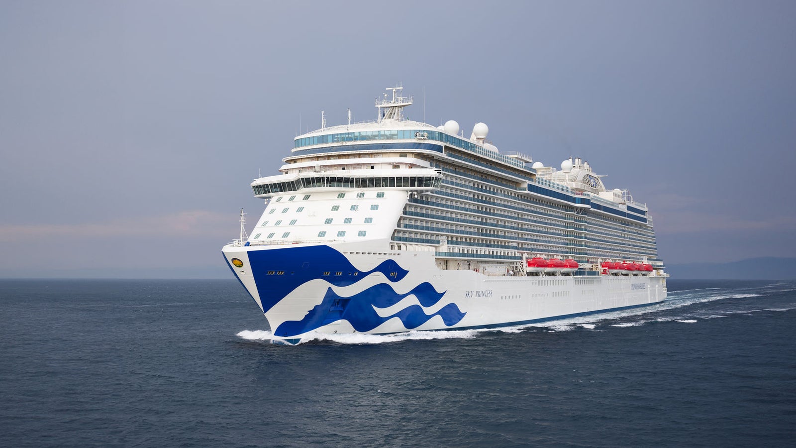 Fincantieri | Gruppo - Business - Prodotti - Navi da Crociera - Sky Princess: Grande nave da crociera di lusso operativa etichettata 'Sky Princess', naviga su acque calme dell'oceano sotto cielo nuvoloso, design elegante con grafica a onda blu sullo scafo, più ponti visibili, scialuppe di salvataggio rosse lungo i lati.