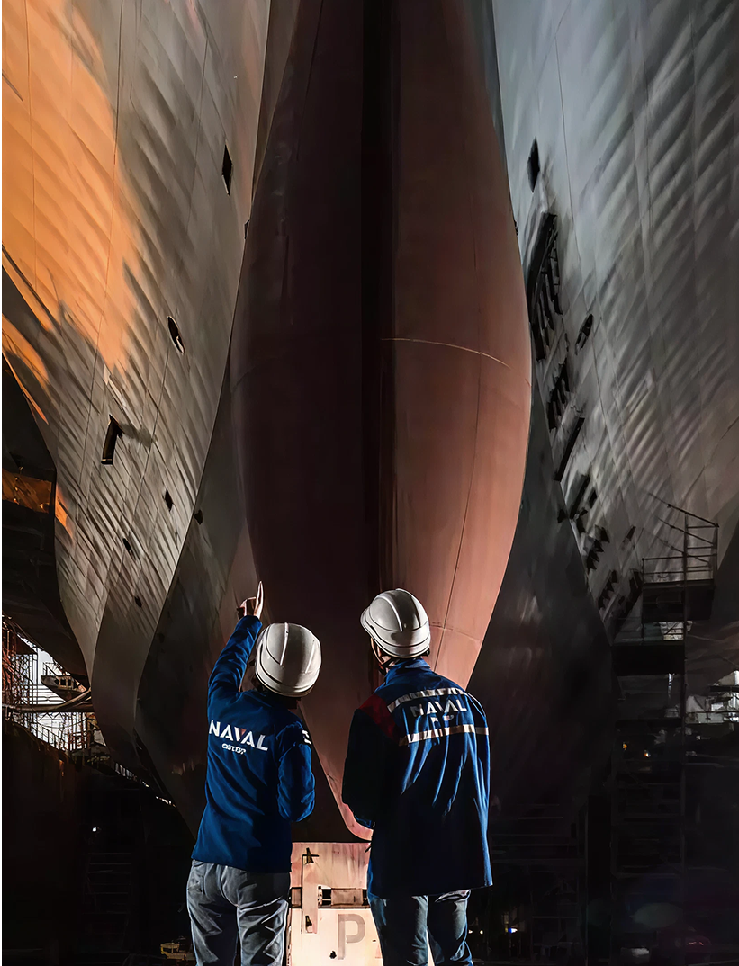Fincantieri | Group - Companies - Subsidiaries and Affiliates - Naviris: Two engineers wearing helmets and blue "Naval" jackets examine the large ship hull in a dimly lit shipyard, industrial scaffolding and shadows highlight the scale, emphasis on technical precision and maritime engineering.