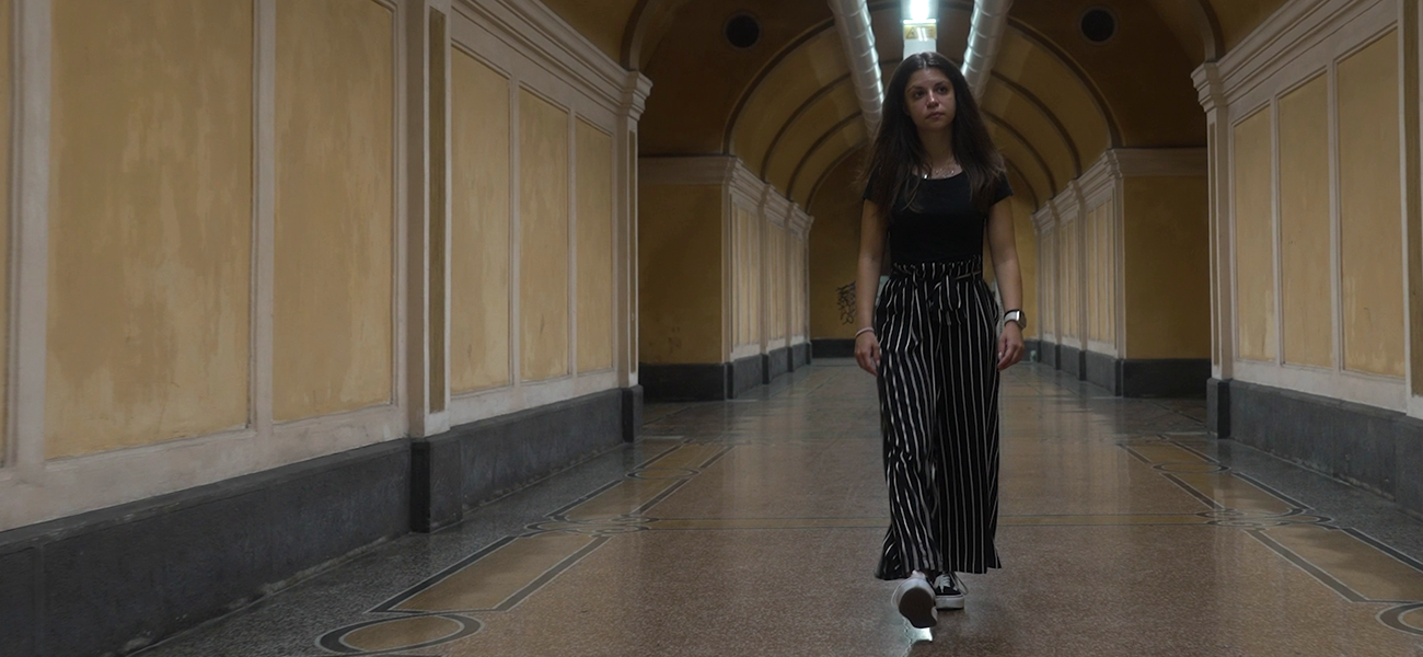 Fincantieri – Persone: Donna vestita con top nero e pantaloni a righe cammina con passo sicuro in un elegante corridoio con soffitti a volta, pareti gialle e dettagli architettonici raffinati, pavimento piastrellato lucido, atmosfera moderna e sofisticata.