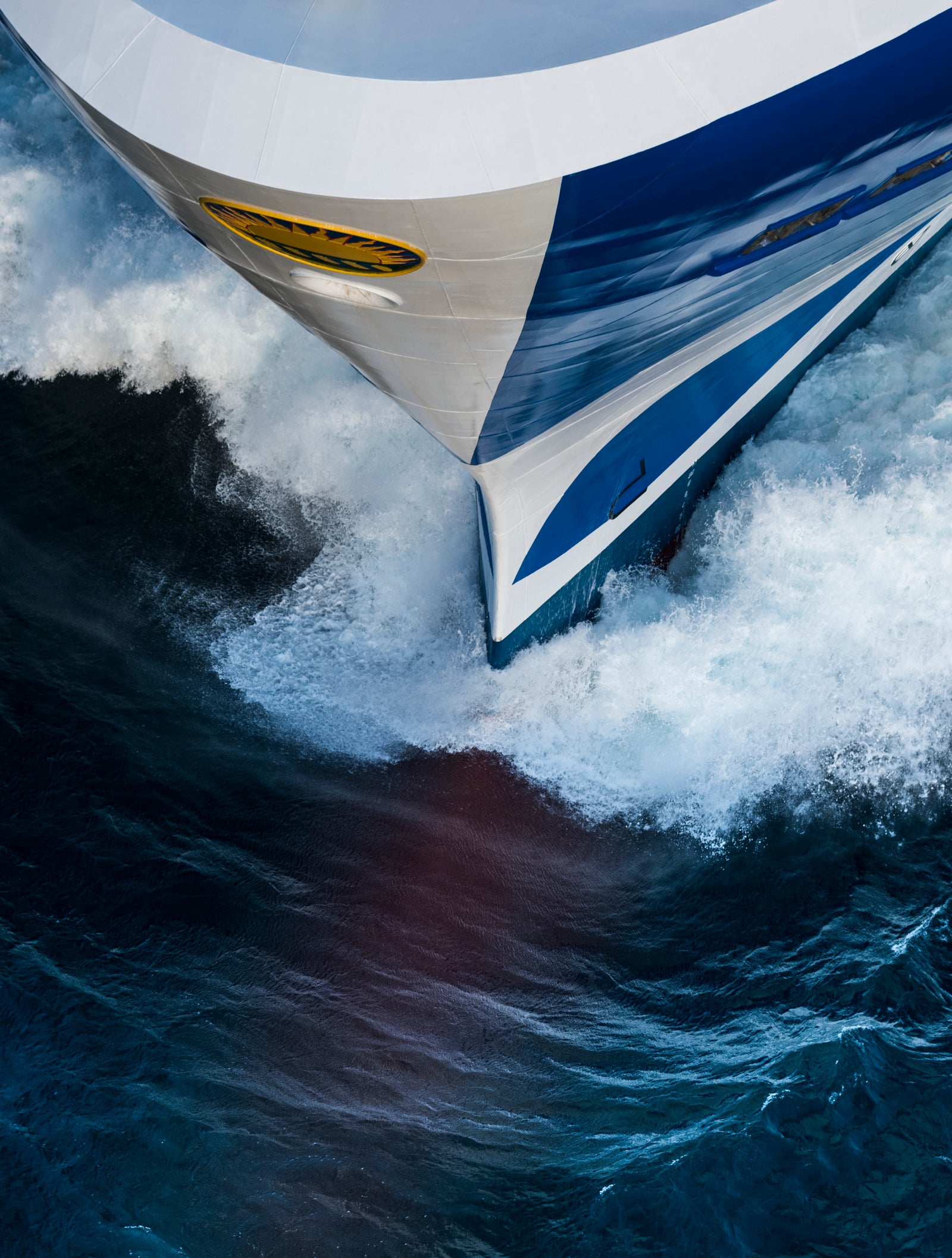 Fincantieri | Group - Business - Products - Cruise Ships - Majestic Princess: Dynamic close-up of Majestic Princess cruise ship bow in operation cutting through ocean waves, white foam and turbulence, elegant blue and white design, visible text on hull.