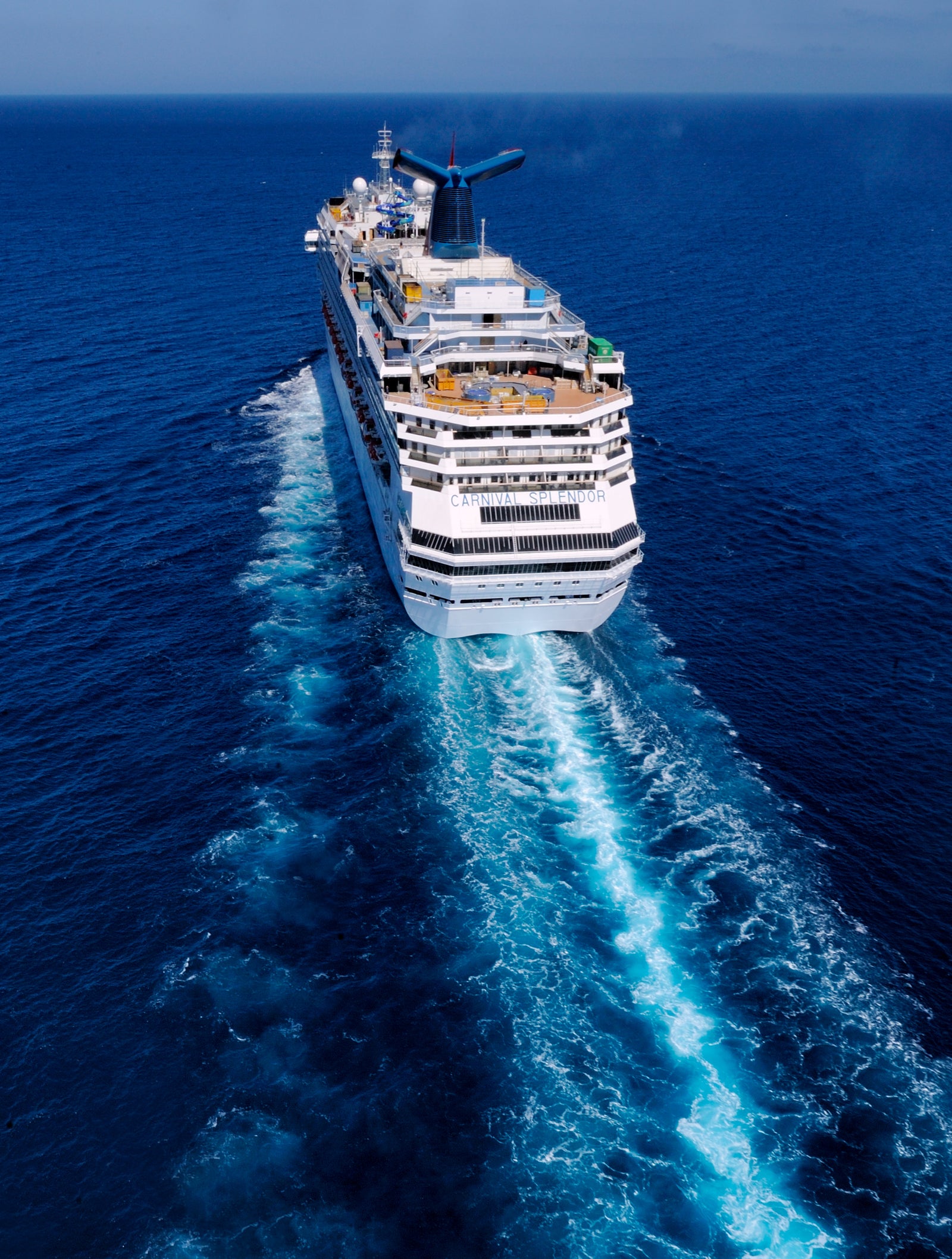 Fincantieri | Group - Business - Products - Cruise Ships - Carnival Splendor operativa: Stern view of large operational cruise ship Carnival Splendor sailing on deep blue waters, prominent white wake behind, multiple decks, lifeboats, distinctive red and blue funnel, vast ocean expanse under clear sky.