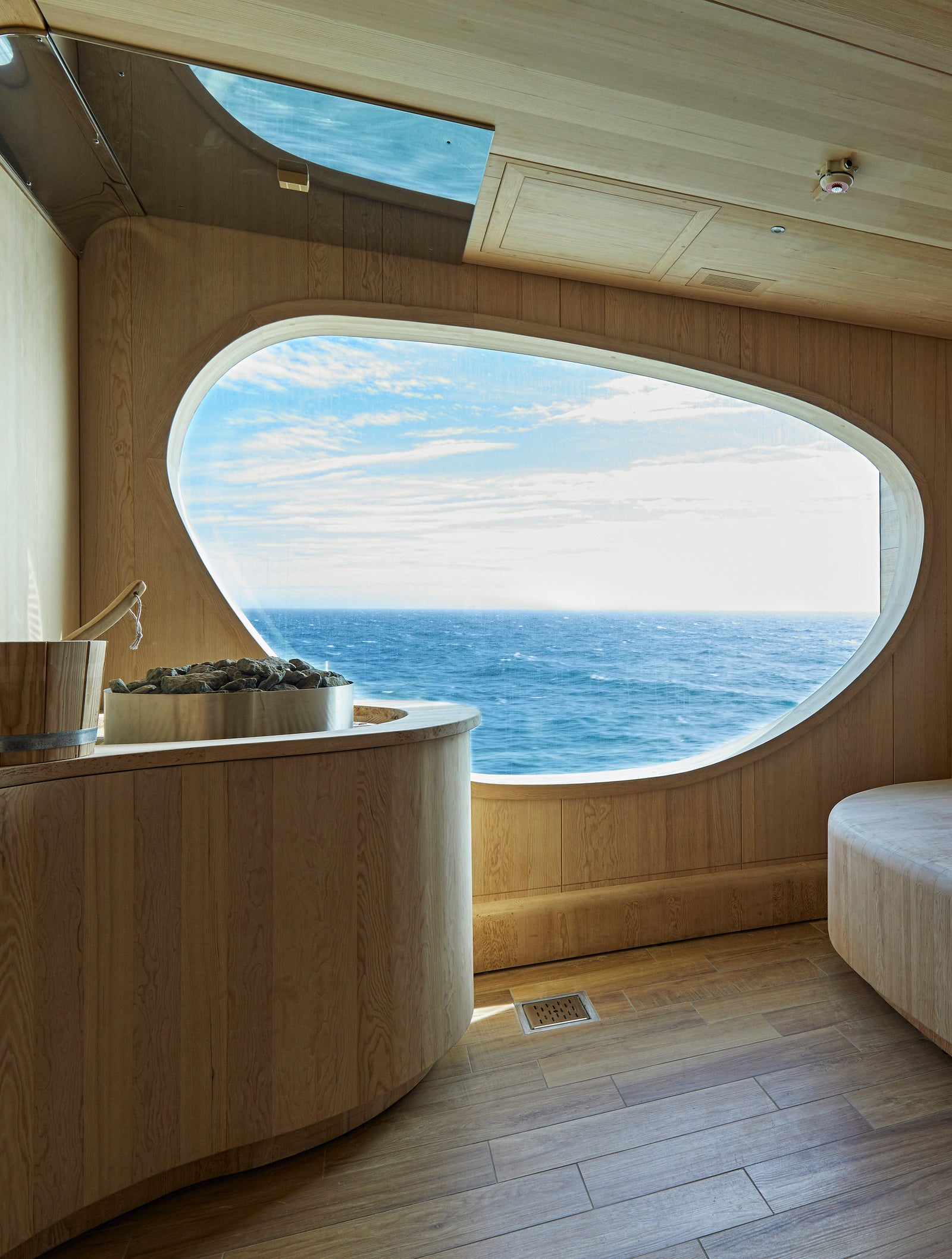 Fincantieri | Gruppo - Business - Prodotti - Navi da Crociera - Le Lapérouse: Interno sauna contemporaneo con sedute curve in legno, grande finestra ovale sul mare, design minimalista con tonalità naturali e linee pulite, secchio e mestolo in legno vicino alle pietre riscaldanti, atmosfera da spa.