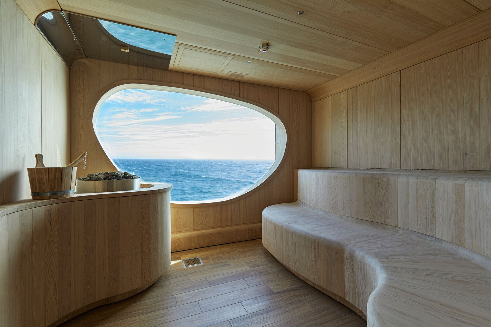 Fincantieri | Gruppo - Business - Prodotti - Navi da Crociera - Le Lapérouse: Interno sauna contemporaneo con sedute curve in legno, grande finestra ovale sul mare, design minimalista con tonalità naturali e linee pulite, secchio e mestolo in legno vicino alle pietre riscaldanti, atmosfera da spa.