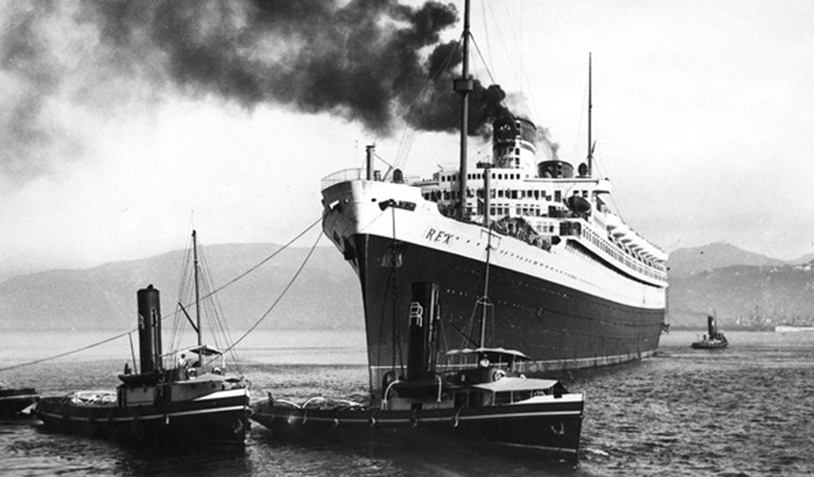 Fincantieri | About Us - Heritage and Future: Large steamship with the name 'REY' assisted by two tugboats in a port, visible smoke, black and white photo with mountainous background and calm waters, historic and industrial maritime atmosphere.