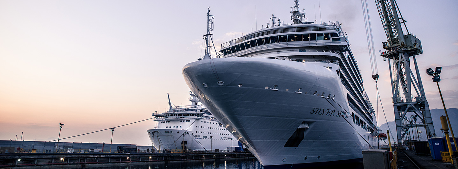 Fincantieri | Group - Business - Products - Systems Integration - Silver Spirit cruise ship: Prominently docked at a harbor during sunset, another cruise ship in the background; industrial cranes and calm waters create a serene maritime setting, ship name 'Silver Spirit' clearly visible on the hull.