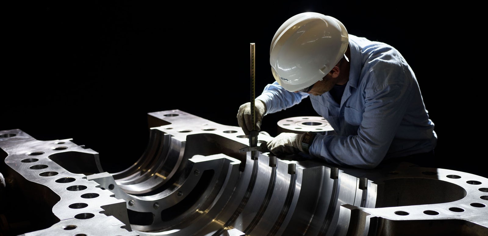 Fincantieri | Group - About Us - Strategic Vision - Business Model: A worker wearing a white safety helmet and gloves inspects a large, intricately machined metal component in a dark industrial setting. The component features multiple circular holes and curved grooves, indicating precision engineering. Lighting highlights the metallic surface and craftsmanship.