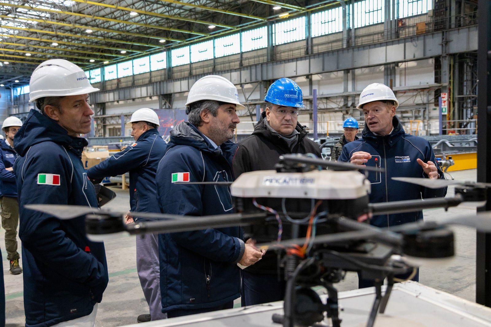 Newsroom - Comunicati stampa - 2025 - Gruppo Fincantieri: Fincantieri: visita del Commissario Europeo Apostolos Tzitzikostas al cantiere di Monfalcone: Delegazione di tecnici e manager Fincantieri con caschi e abbigliamento aziendale insieme al Commissario Europeo Apostolos Tzitzikostas, visita al cantiere di Monfalcone con drone in primo piano e officina navale sullo sfondo.