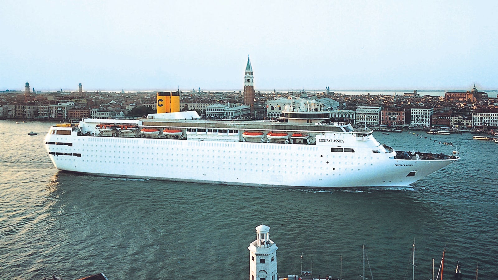 Fincantieri | Group - Business - Products - Cruise Ships - Costa Classica: Side view of Costa Classica cruise ship in operation near Venice, white hull, yellow funnel with visible C logo, background with St. Mark’s Campanile, marina, and historic architecture.