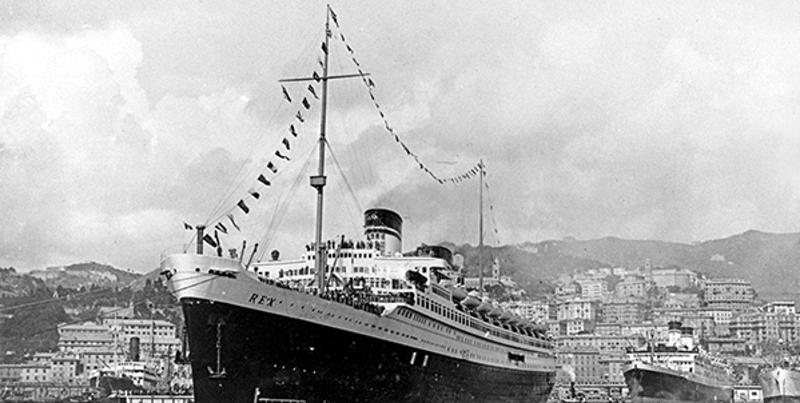 Fincantieri | About Us - Historic Ships: Large ocean liner 'REX' docked in a crowded port, surrounded by smaller boats, coastal city with hills in the background, flags and celebratory banners on the ship.