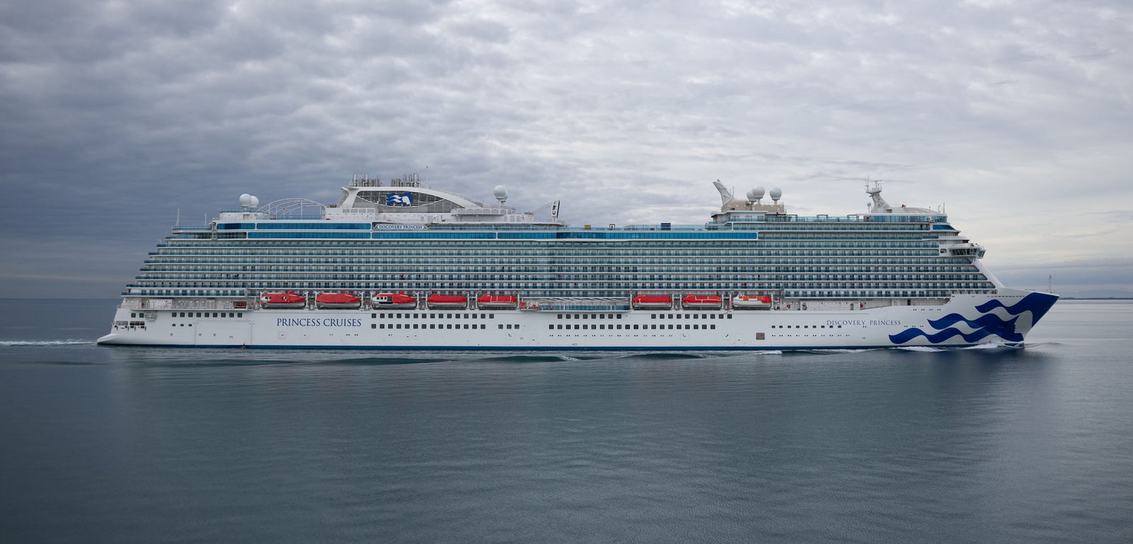 Fincantieri | Group - Business - Products - Cruise Ships - Discovery Princess: Close side view of Discovery Princess Princess Cruises ship in operation, sailing on calm open waters under cloudy sky, multiple decks, red lifeboats, blue wave logo on hull.