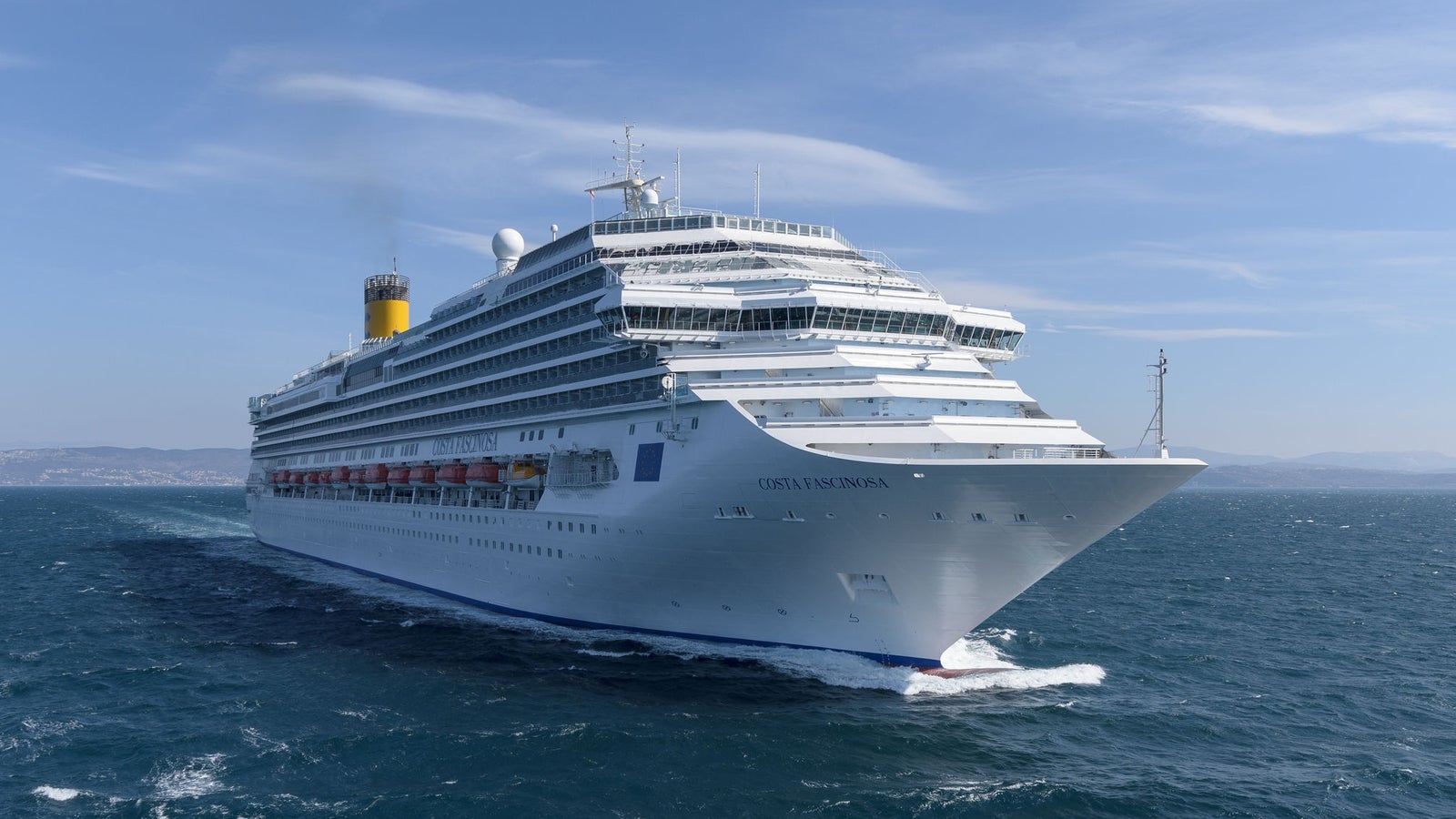 Fincantieri | Group - Business - Products - Cruise Ships - Costa Fascinosa: Side view of Costa Fascinosa cruise ship in operation, white hull with multiple passenger decks, visible lifeboats and identifying Costa Fascinosa name on hull.