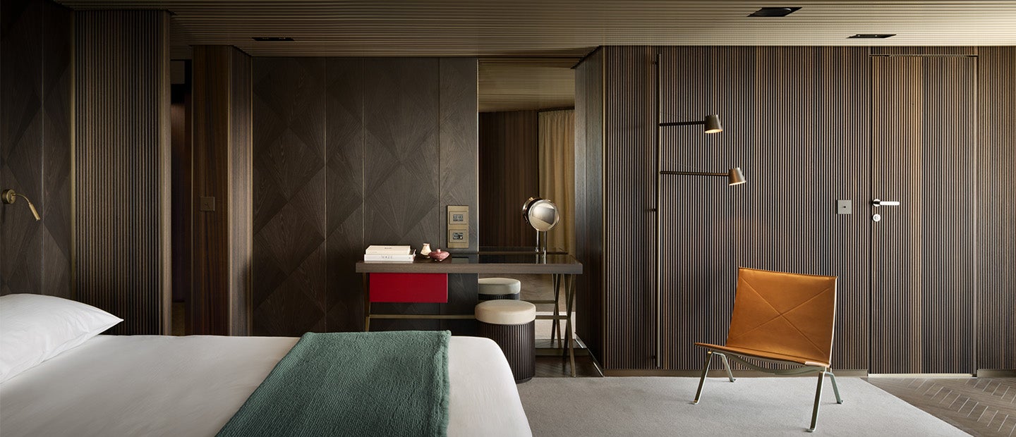  Fincantieri | Gruppo - Società - Controllate e collegate - Operae Interiors: Camera d’hotel contemporanea con design minimalista, pareti in legno e palette neutra; letto ordinato, scrivania con specchio tondo e poltrona in pelle, decorazione con linee pulite e illuminazione moderna per atmosfera sofisticata.