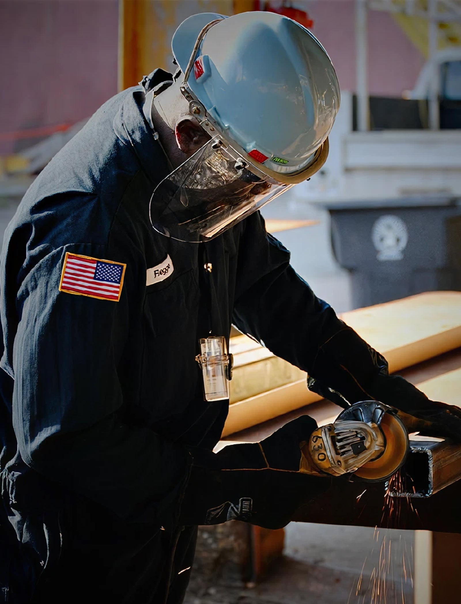 Fincantieri | Gruppo - Società - Controllate e collegate - Marine Systems North America: Operaio con dispositivi di protezione lavora con smerigliatrice manuale su superficie metallica in ambiente industriale; uniforme scura con patch bandiera USA, guanti e casco, scintille visibili durante il taglio. Sfondo con strutture industriali e cassonetto con simbolo riciclo.