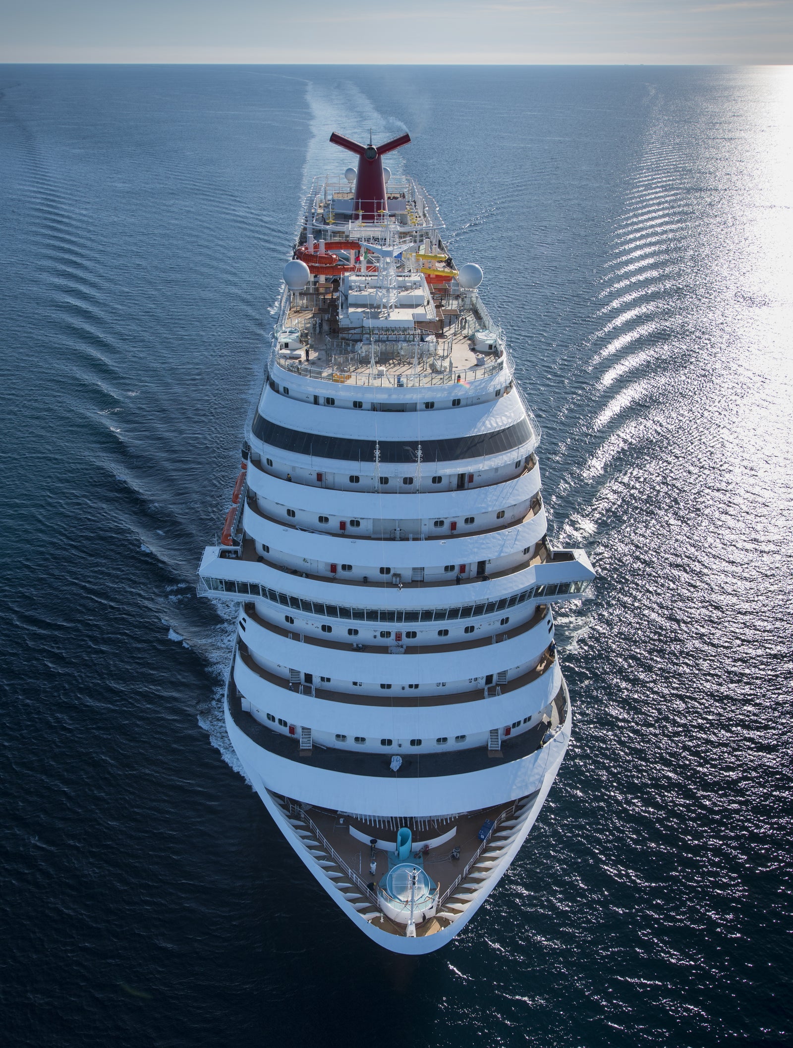 Fincantieri | Group - Business - Products - Cruise Ships - Carnival Vista: Aerial view of Carnival Vista cruise ship in operation, sailing through calm open waters, multiple decks and distinctive red funnel, visible passengers enjoying amenities, serene ocean setting with clear skies.