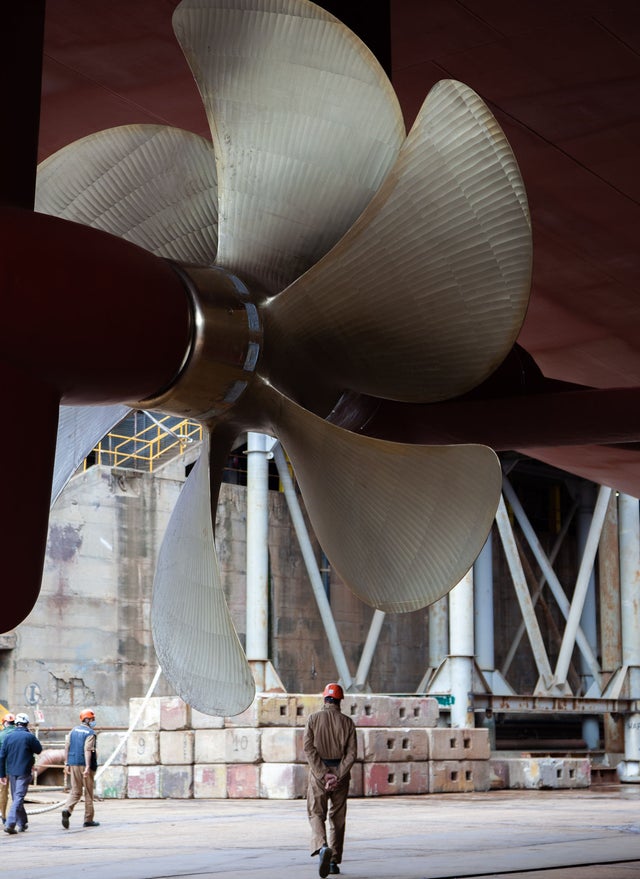 Fincantieri | Group - Business - Products - Stabilization, Propulsion, Positioning, and Generation Systems - Ship propeller: Large ship propeller prominently displayed in the foreground; workers and industrial equipment visible in background at shipyard or dock.