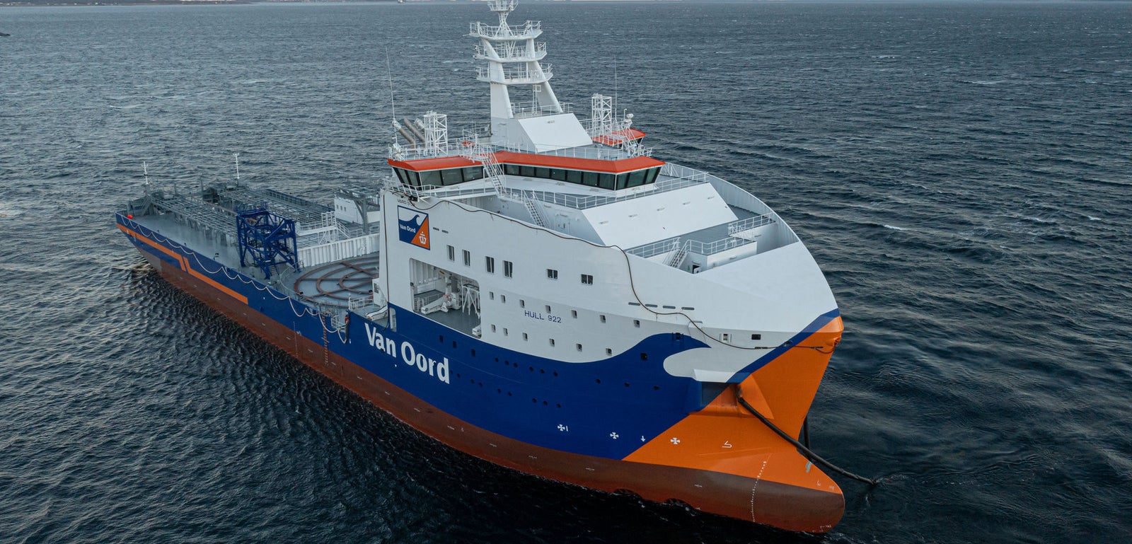 Prodotto - Nave Van Oord Calypso: Vista laterale della nave operativa in navigazione al tramonto, scafo arancione e blu, testo Van Oord chiaramente visibile, sfondo con montagne lontane e orizzonte marino.