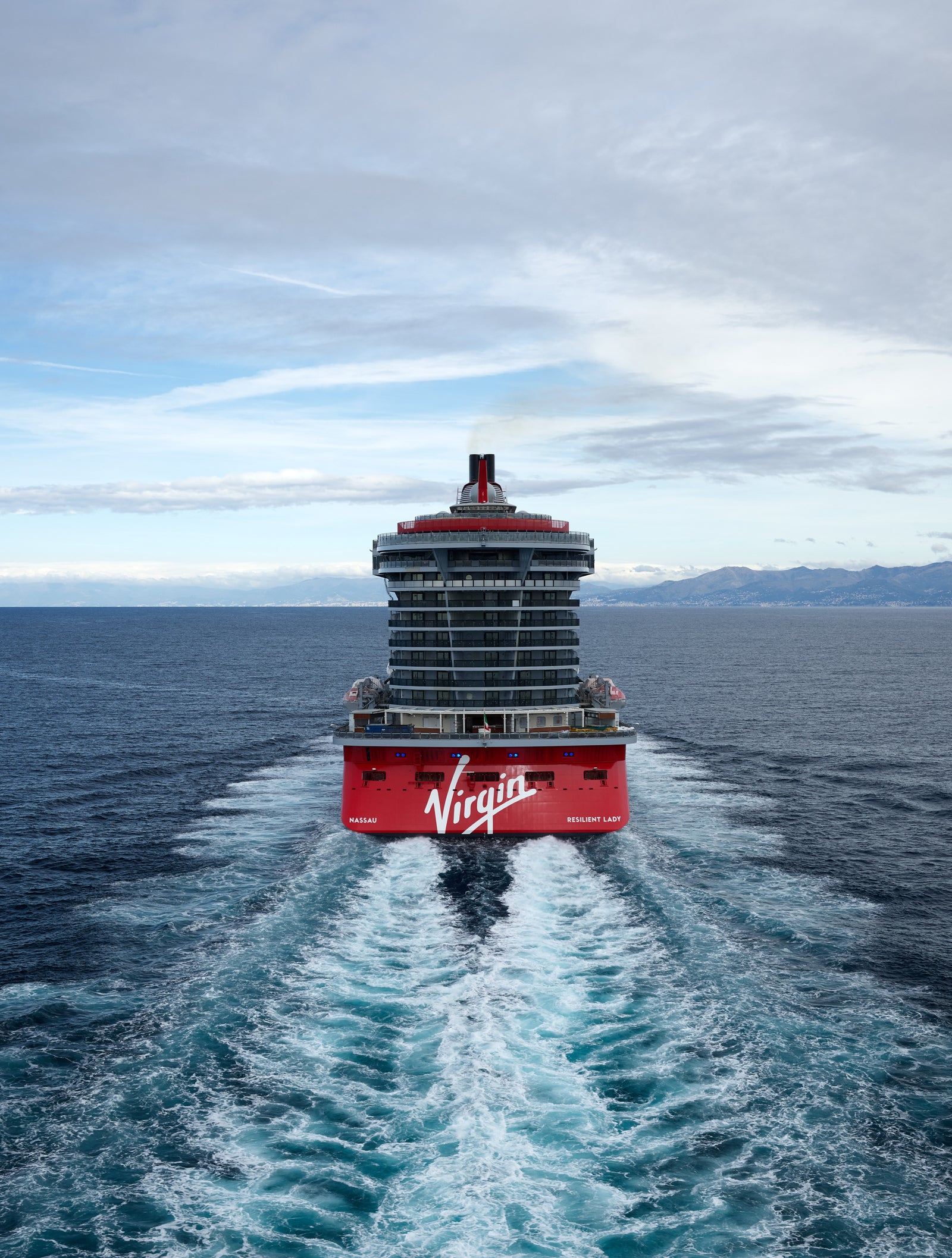 Fincantieri | Group - Business - Products - Cruise Ships - Resilient Lady: Large operational cruise ship with modern design, distinctive red funnel, multiple decks, white wave wake, sailing on calm waters under partly cloudy sky, wide maritime setting with distant mountains on horizon.