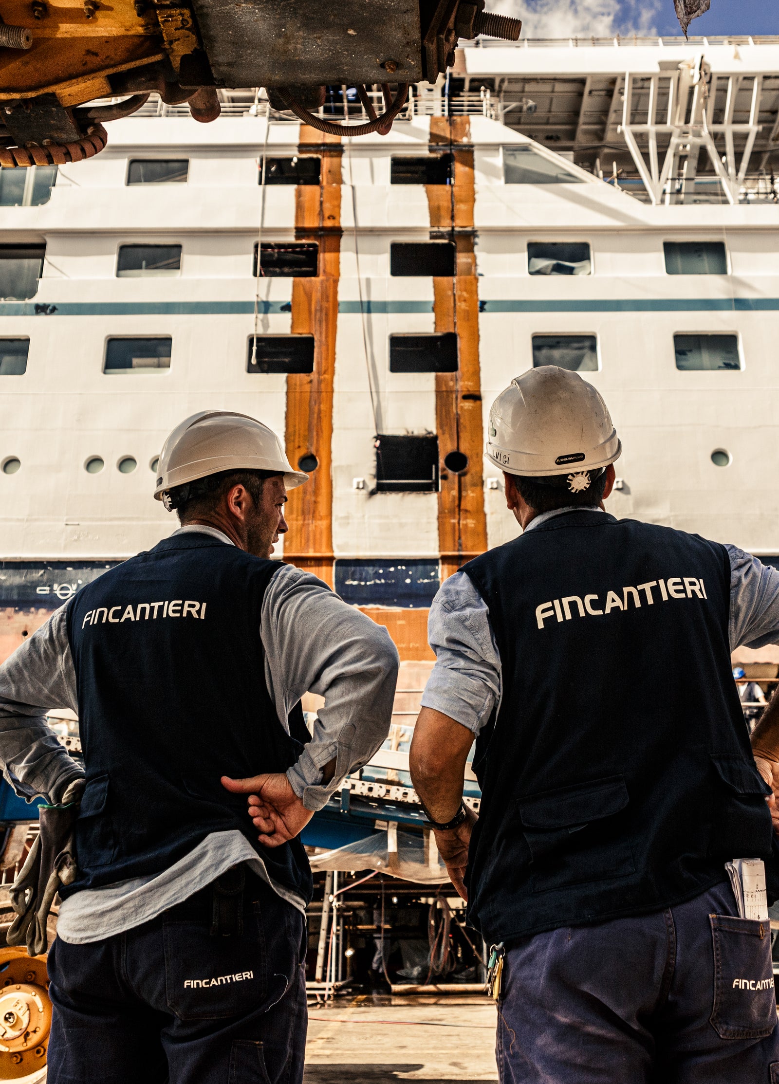 Fincantieri Group | About Us: Two operators wearing Fincantieri helmets and vests observe a large cruise ship in a shipyard, with industrial equipment and scaffolding in the background; hull and upper decks are visible, with bright daylight highlighting the scene.