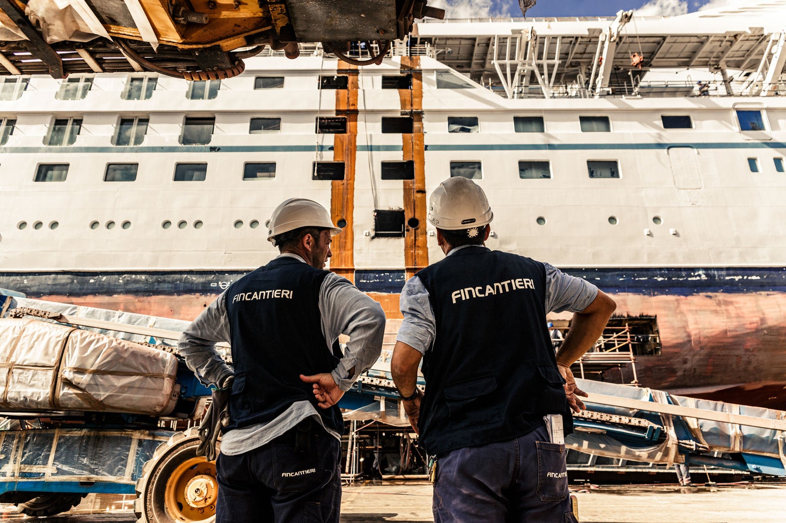 Gruppo Fincantieri | Chi siamo: Due operatori con caschi e gilet Fincantieri osservano una grande nave da crociera in cantiere navale, sfondo con attrezzature industriali e impalcature, scafo e ponti superiori visibili, luce diurna intensa che mette in risalto la scena.