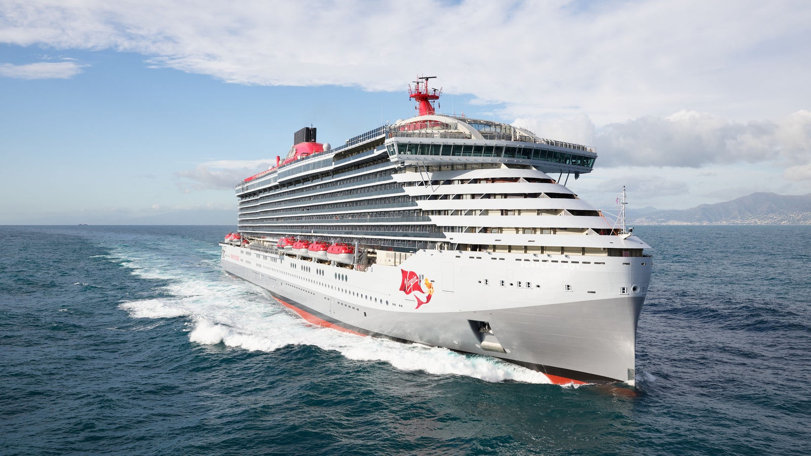 Fincantieri | Group - Business - Products - Cruise Ships - Scarlet Lady: Large operational cruise ship, white with red details and red mermaid logo, sailing on open waters with visible wakes, partly cloudy sky and distant mountainous coastline in background.