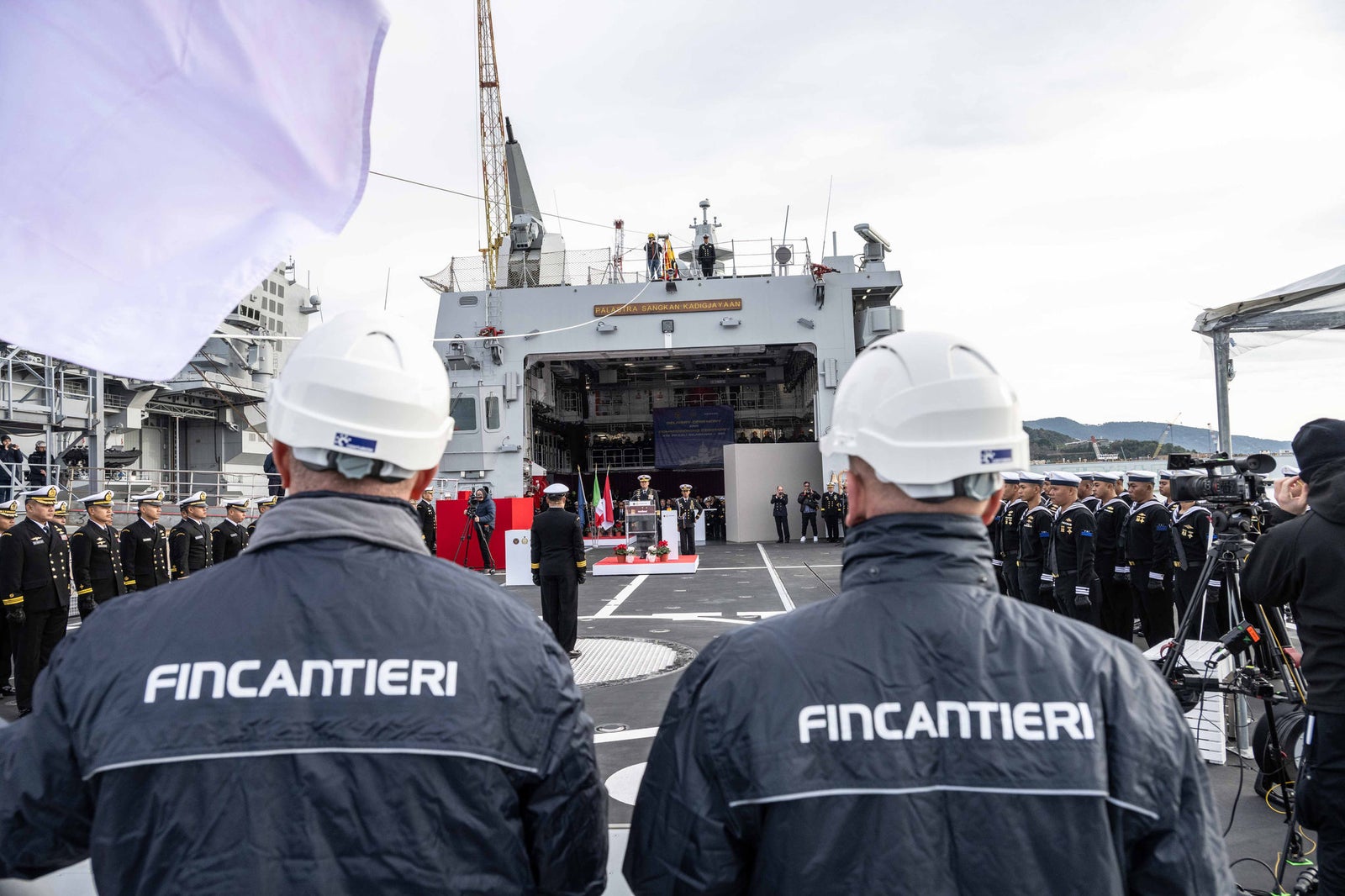 newsroom - press releases - 2025 - fincantieri group: fincantieri delivers the ppa "kri prabu siliwangi-321" to the indonesian navy: two fincantieri workers in the foreground with helmets, official ceremony on the deck of the "kri prabu siliwangi-321" with officers and crew in uniform, flags and photographers.