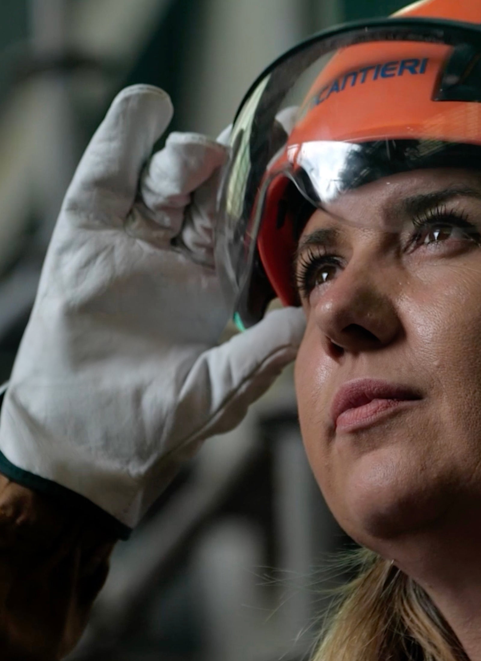 Fincantieri | Group - Sustainability - People and Community: Woman wearing a safety helmet and gloves adjusting her protective glasses, with a determined expression looking upward.