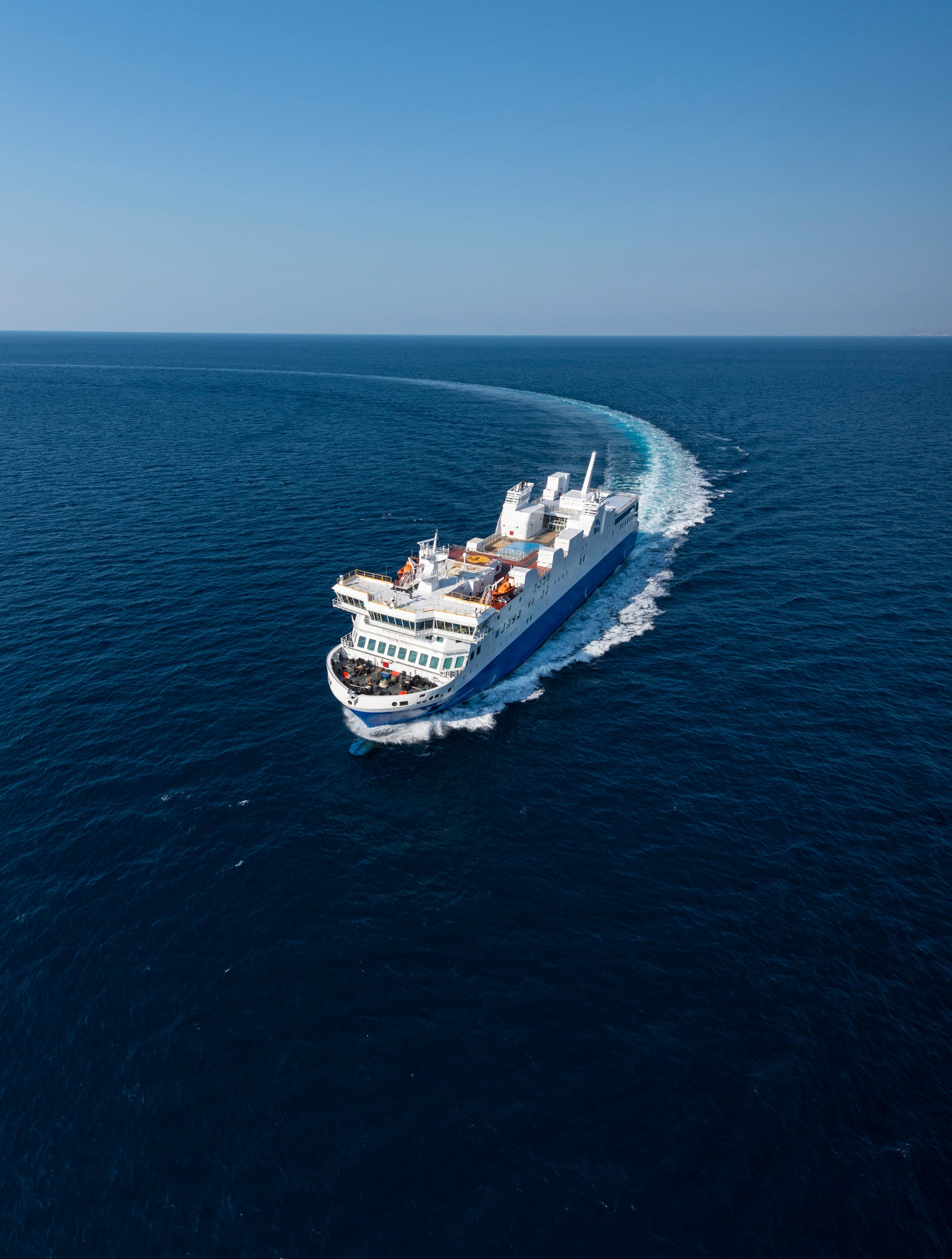 A large white and blue ferry navigates through the open ocean under a clear blue sky. The vessel creates a visible wake as it moves forward, showcasing its size and power. The serene setting highlights the vastness of the sea and the tranquility of the journey.