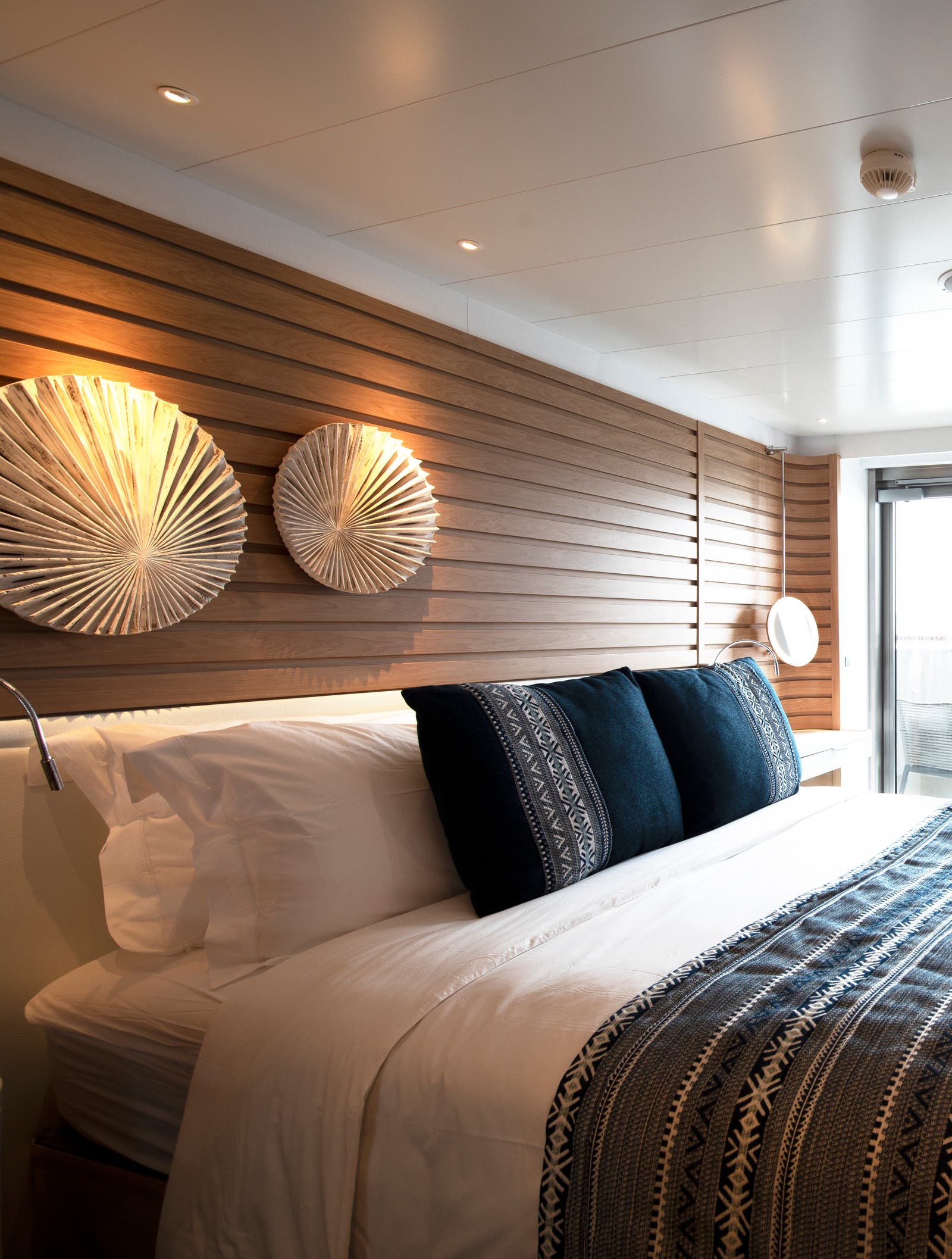Fincantieri | Gruppo - Business - Prodotti - Navi da Crociera - Le Lapérouse: Cabina elegante e moderna con letto confortevole, biancheria decorata, cuscini ornamentali, pannelli in legno, illuminazione ambientale, specchio circolare, balcone con porta vetrata che offre vista mare e luce naturale, atmosfera serena.