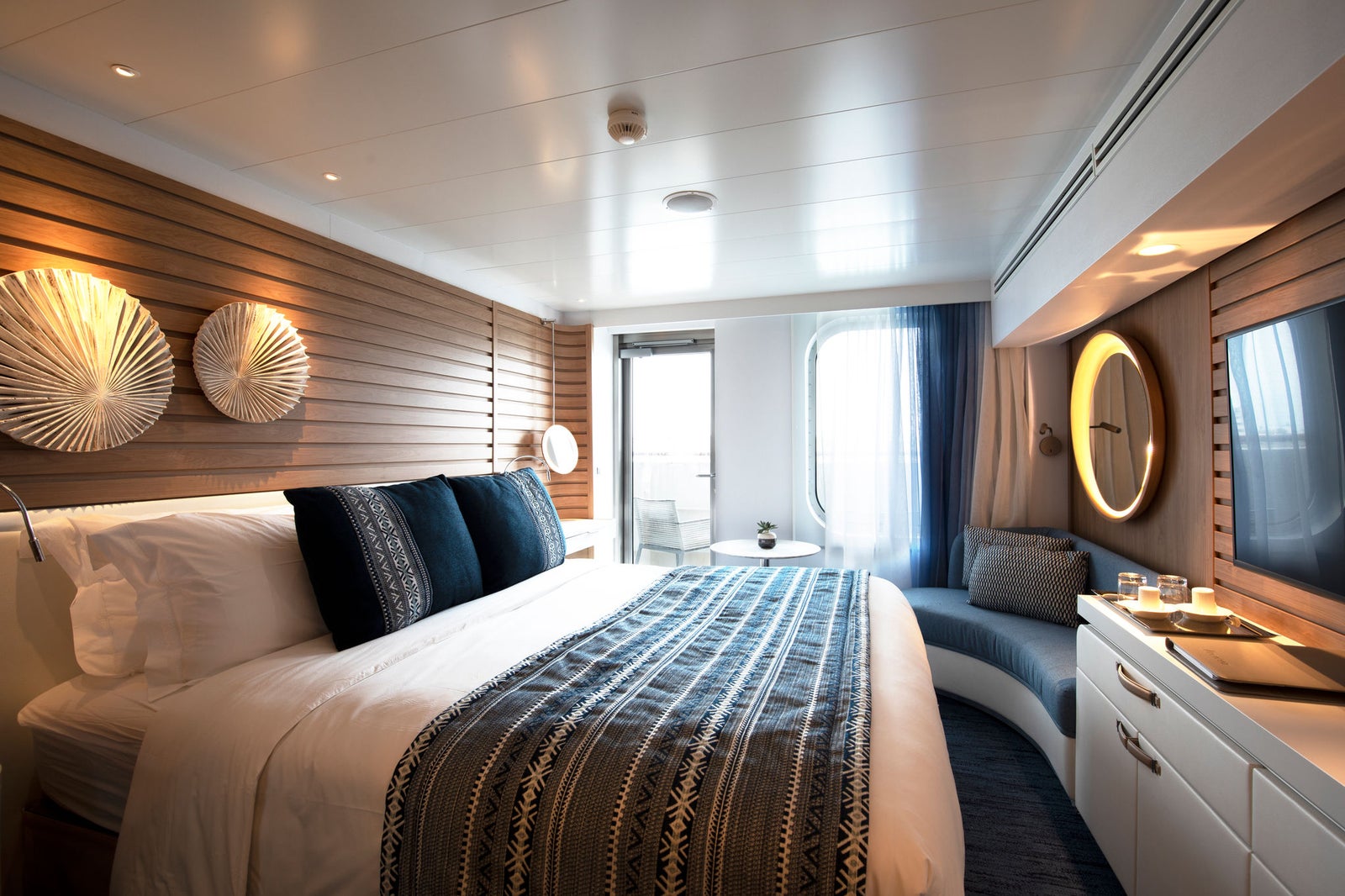 Fincantieri | Gruppo - Business - Prodotti - Navi da Crociera - Le Lapérouse: Cabina elegante e moderna con letto confortevole, biancheria decorata, cuscini ornamentali, pannelli in legno, illuminazione ambientale, specchio circolare, balcone con porta vetrata che offre vista mare e luce naturale, atmosfera serena.