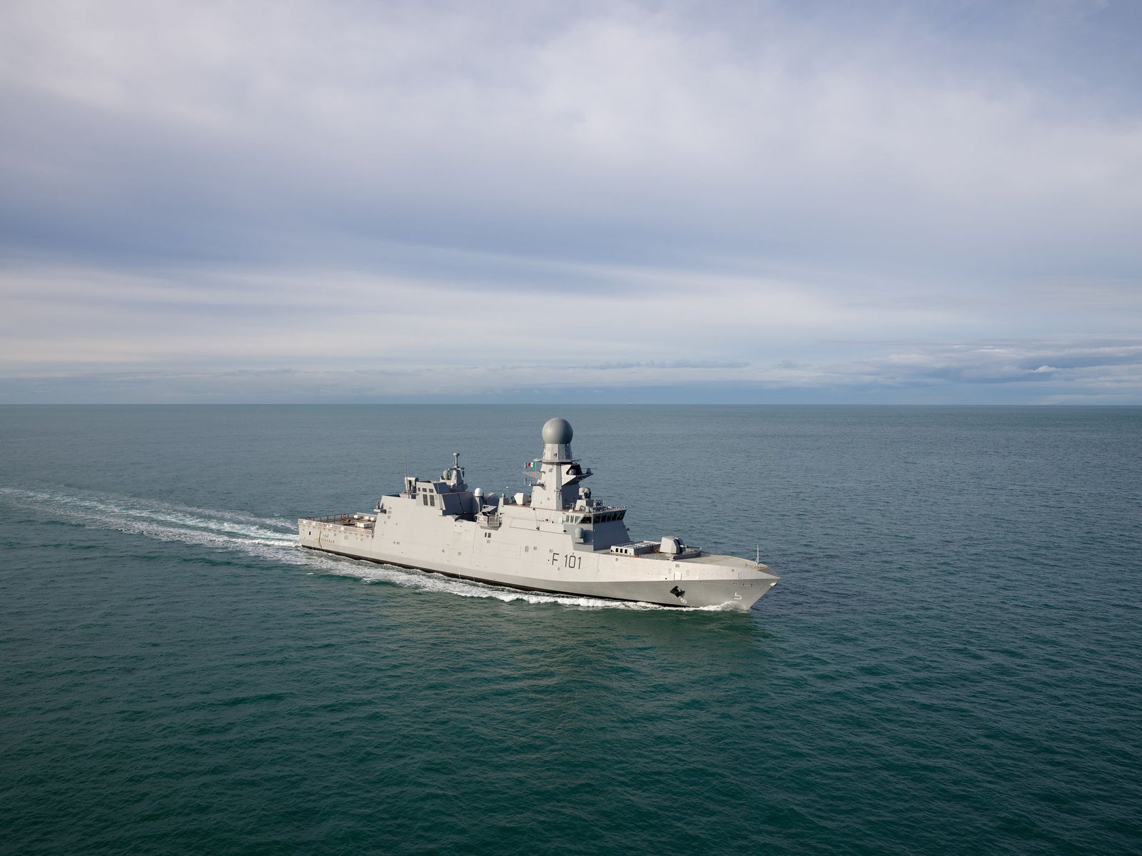 Fincantieri | Group - Companies - Subsidiaries and Affiliates - Naviris: Operational military ship 'F 10 1' sailing on calm waters under a cloudy sky, hull in neutral tones equipped with advanced radars on the deck; open sea to the horizon, serene yet powerful view.