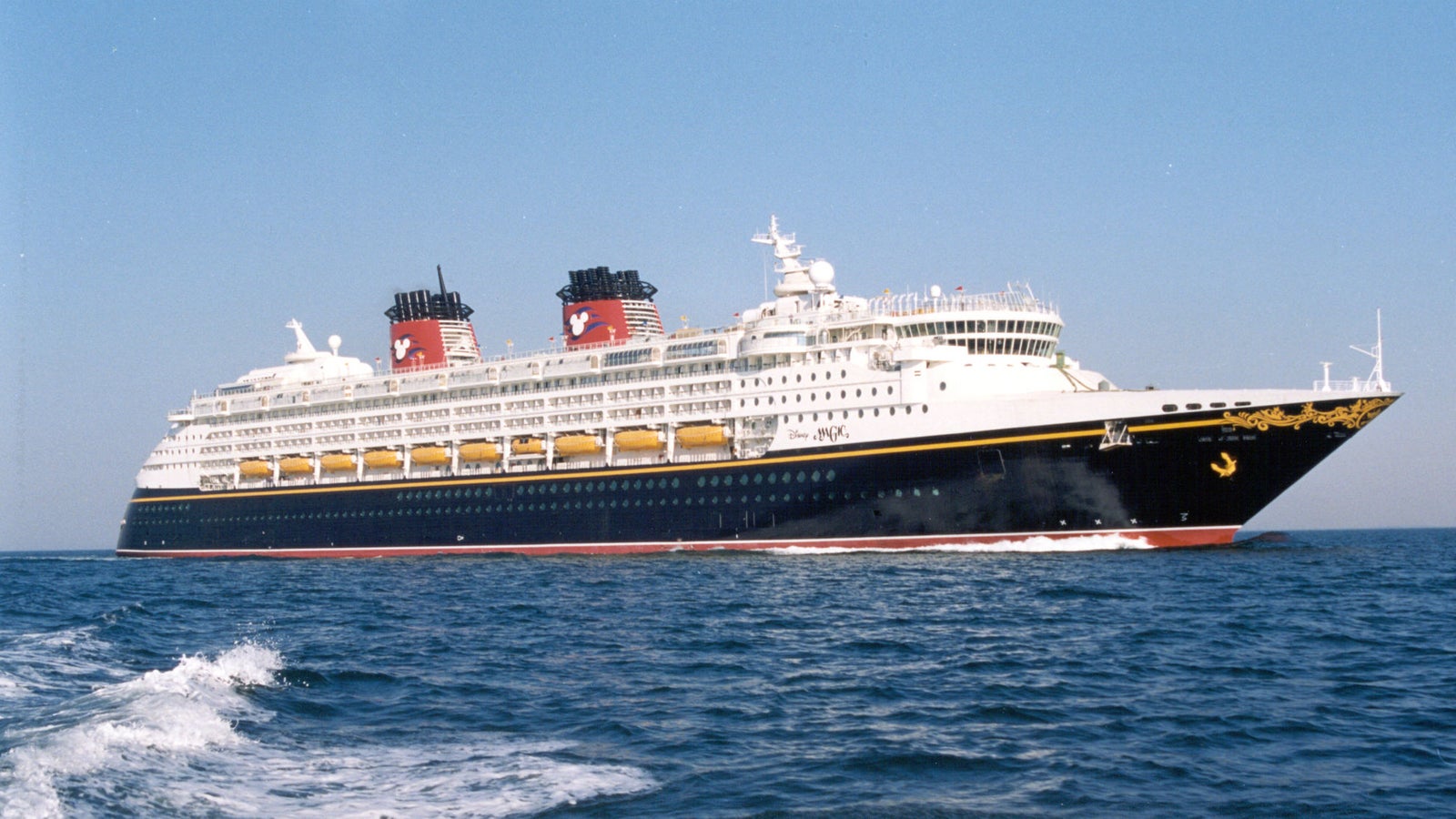 Fincantieri | Group - Business - Products - Cruise Ships - Disney Magic: Side view of Disney Magic cruise ship in operation navigating, distinctive dual red funnels, multiple decks, visible yellow lifeboats, sleek design, visible wake.