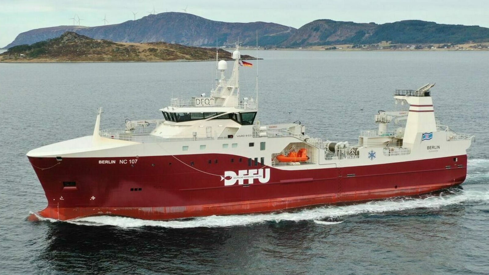 Prodotto - Peschereccio Berlin: Vista laterale della nave operativa in navigazione vicino a un’isola costiera, scafo rosso con scritta SPFU, sovrastruttura bianca, sfondo con colline distanti e mare calmo.