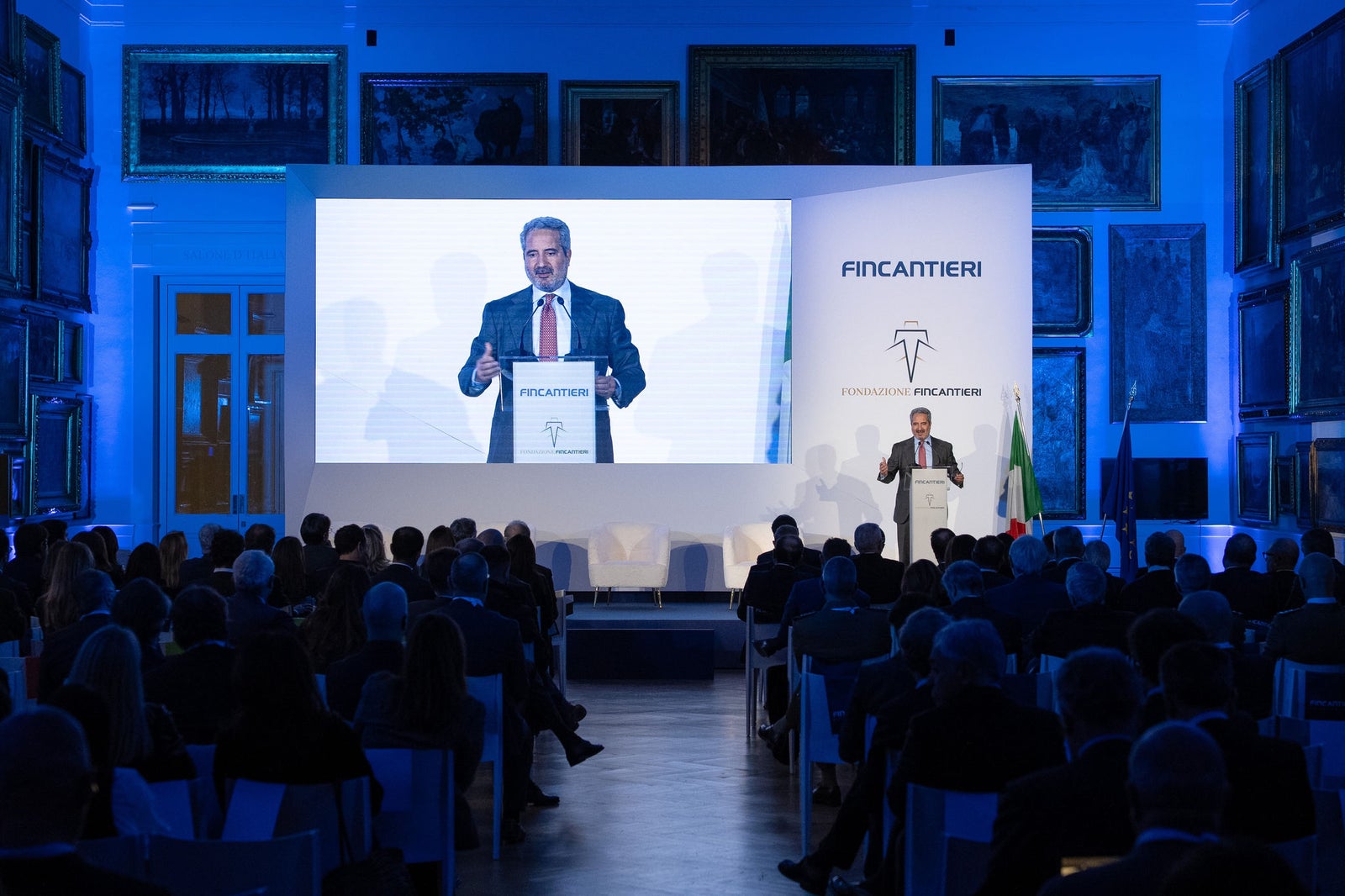 Newsroom - Comunicati stampa - 2025 - Gruppo Fincantieri: Un anno di Fondazione Fincantieri: storia, innovazione e impatto sociale: Speaker istituzionale alla cerimonia annuale della Fondazione Fincantieri, con platea e palco allestito, maxi schermo, loghi Fondazione e Fincantieri, bandiere italiana ed europea, sala con illuminazione blu e quadri storici alle pareti.