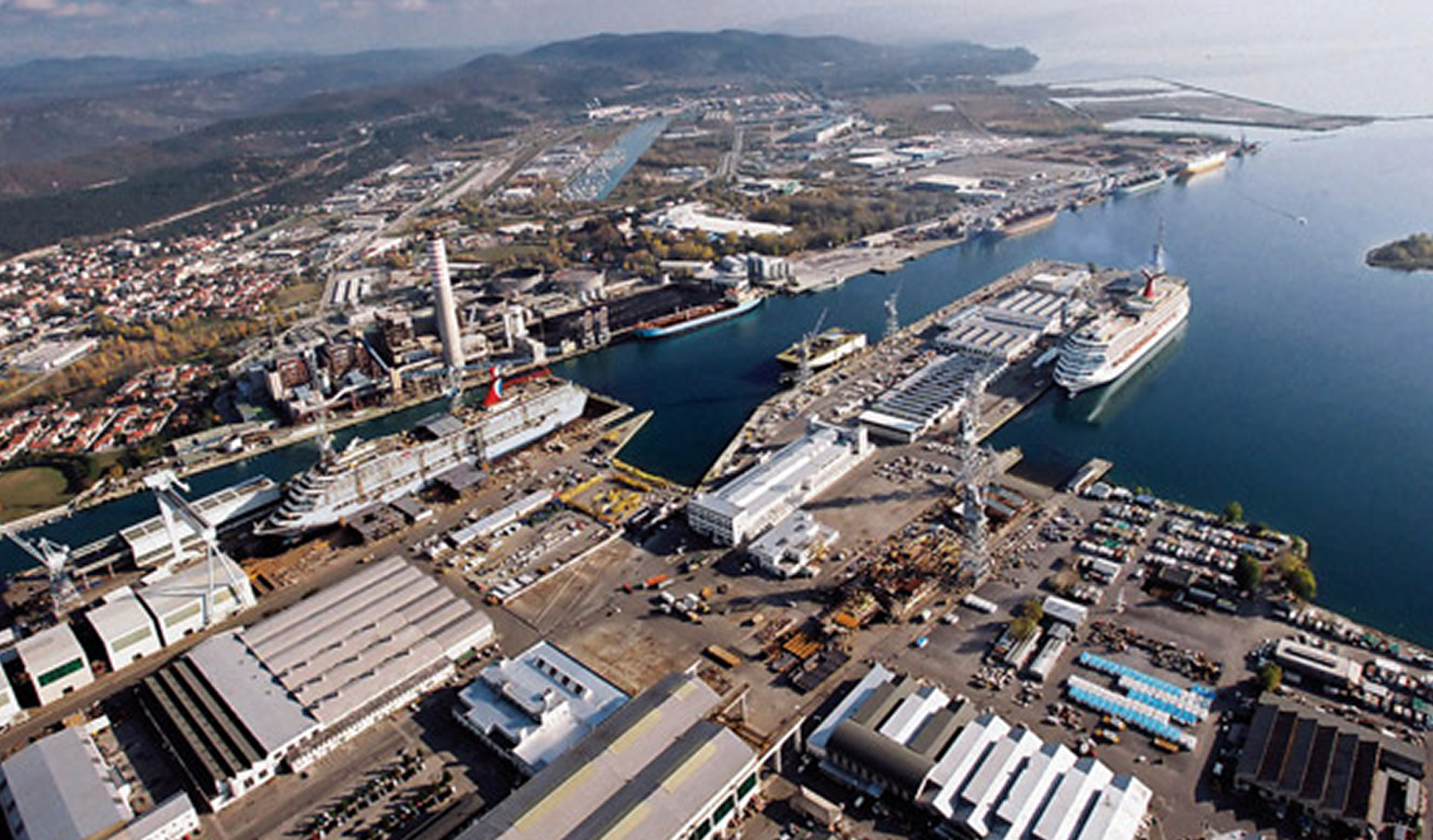Fincantieri | About Us - Heritage and Future: Wide aerial view of an industrial port with docked cruise ships and cargo facilities, warehouses, cranes and surrounding infrastructure, hilly background and water, clear day highlighting maritime activity.