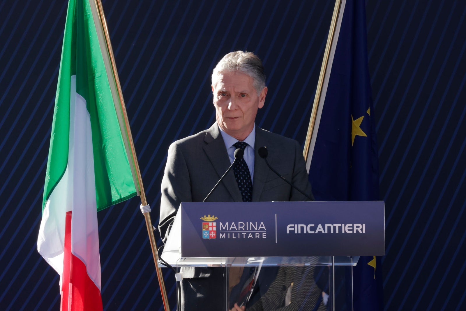 Newsroom - Press releases - 2026 - Fincantieri Group: Launch of the Italian Navy's "Quirinale" major hydro-oceanographic ship: fincantieri representative in suit speaks at podium with marina militare and european flags during launch event.
