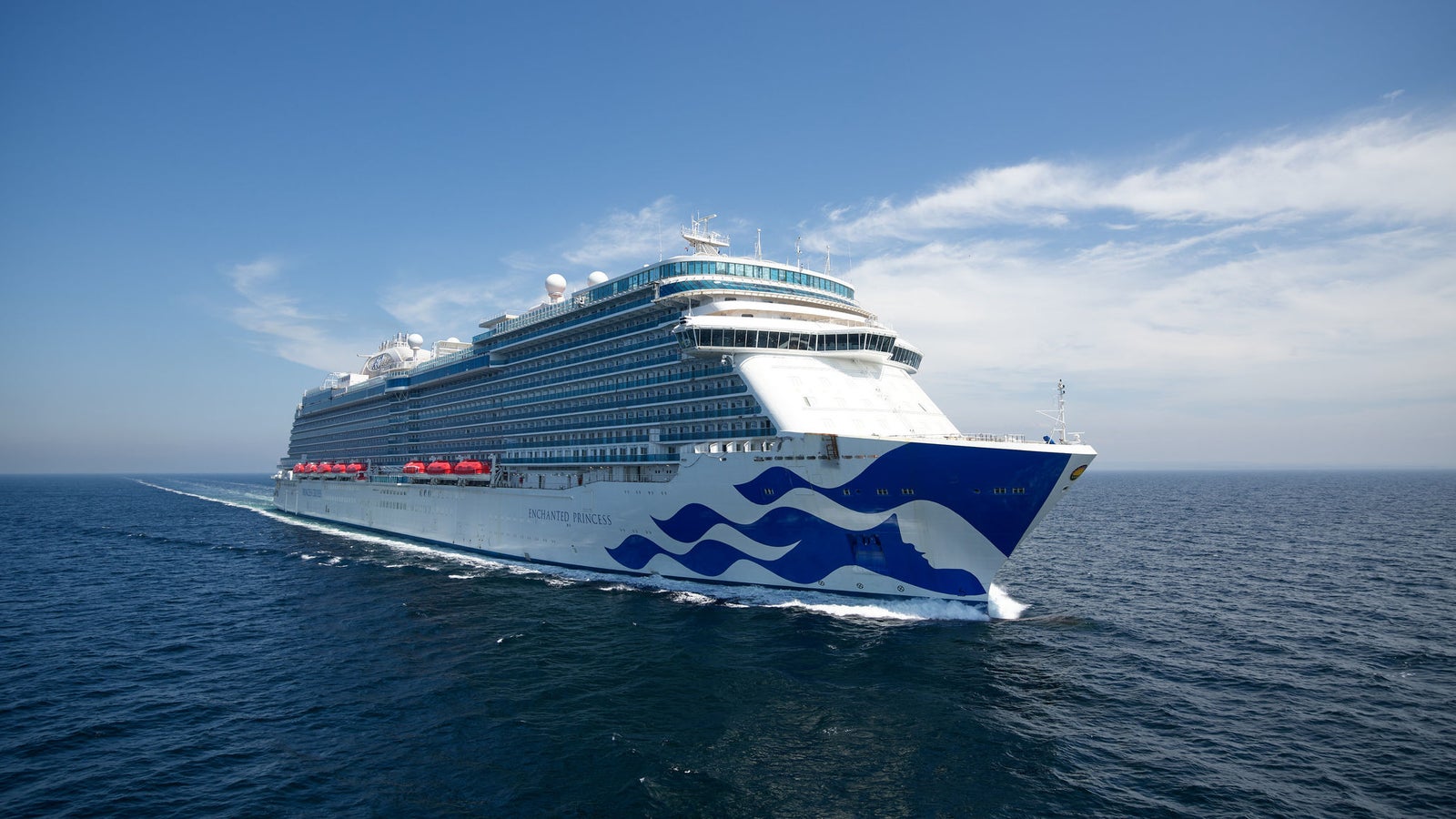 Fincantieri | Group - Business - Products - Cruise Ships - Enchanted Princess: Overhead view of Enchanted Princess cruise ship in operation, sailing on calm open waters under clear blue sky, white and blue exterior with wave patterns on hull, visible lifeboats along sides, upper decks show antennas and recreation areas.