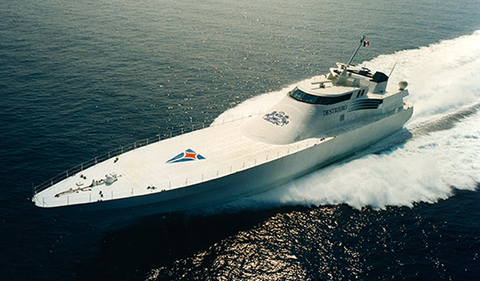 Fincantieri | About Us - Heritage and Future: High-speed yacht with modern design, featuring the name 'DESTIERO' and a geometric logo on the deck, sailing on the open ocean, dynamic movement through the waves under a clear sky.