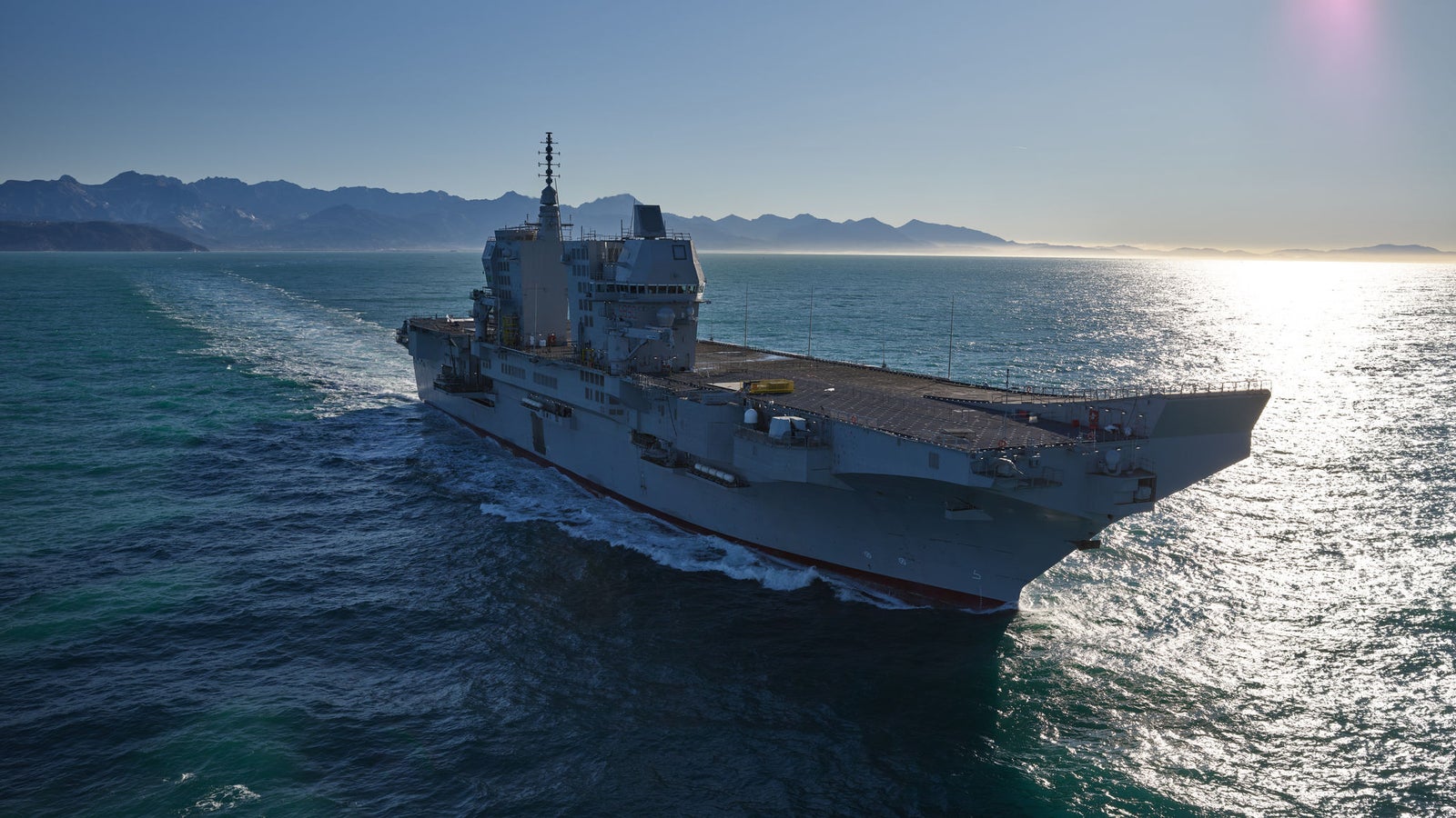 Fincantieri | Group - Business - Products - Naval Vessels - Aircraft Carriers and Amphibious Ships - Trieste: Aircraft carrier "Trieste" in operation, sailing on ocean, mountains in background, sunlight reflecting on water.