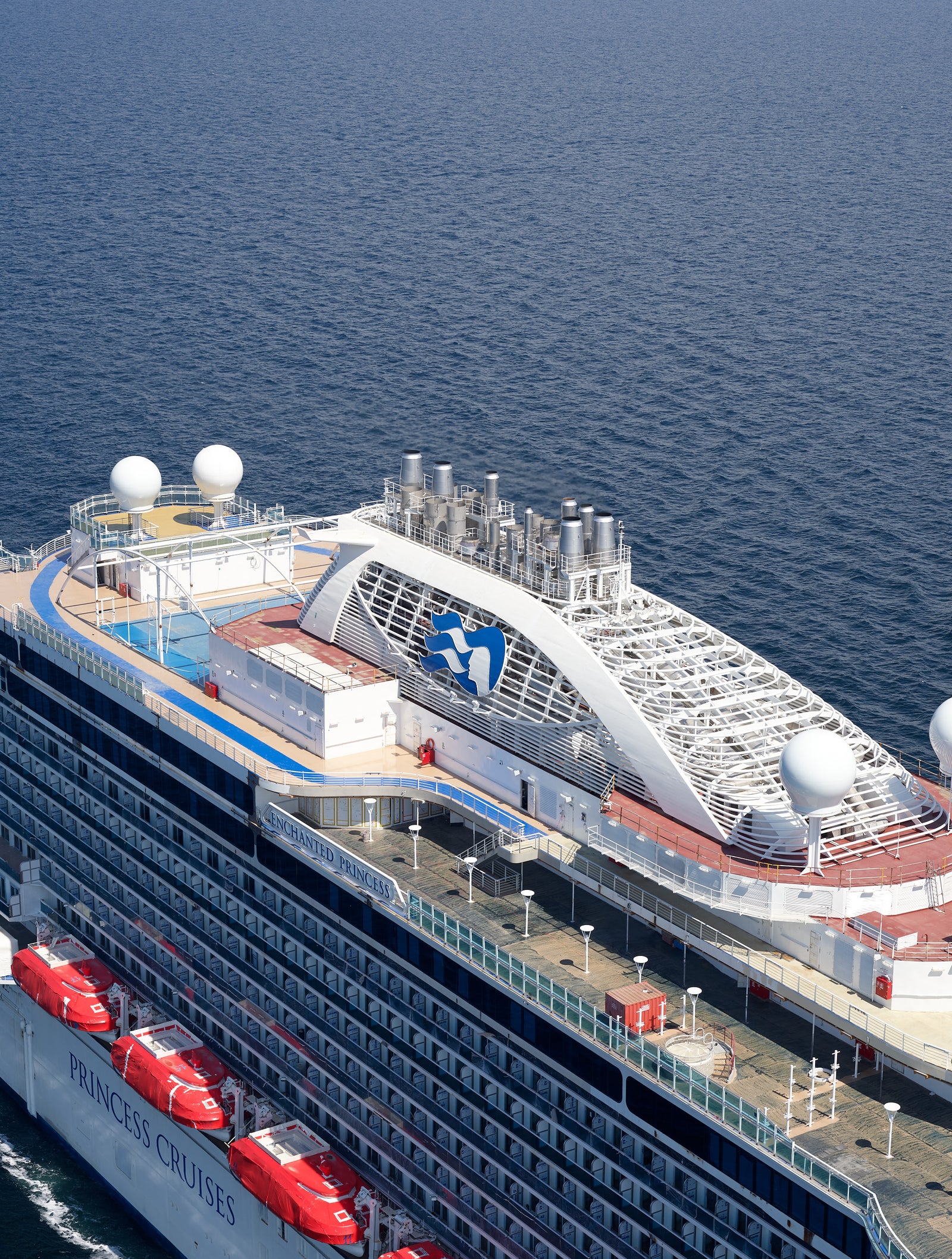 Fincantieri | Group - Business - Products - Cruise Ships - Enchanted Princess: Large operational cruise ship sailing on calm open waters under clear blue sky, white and blue exterior with wave patterns on hull, visible lifeboats along sides, upper decks with antennas and recreation areas.