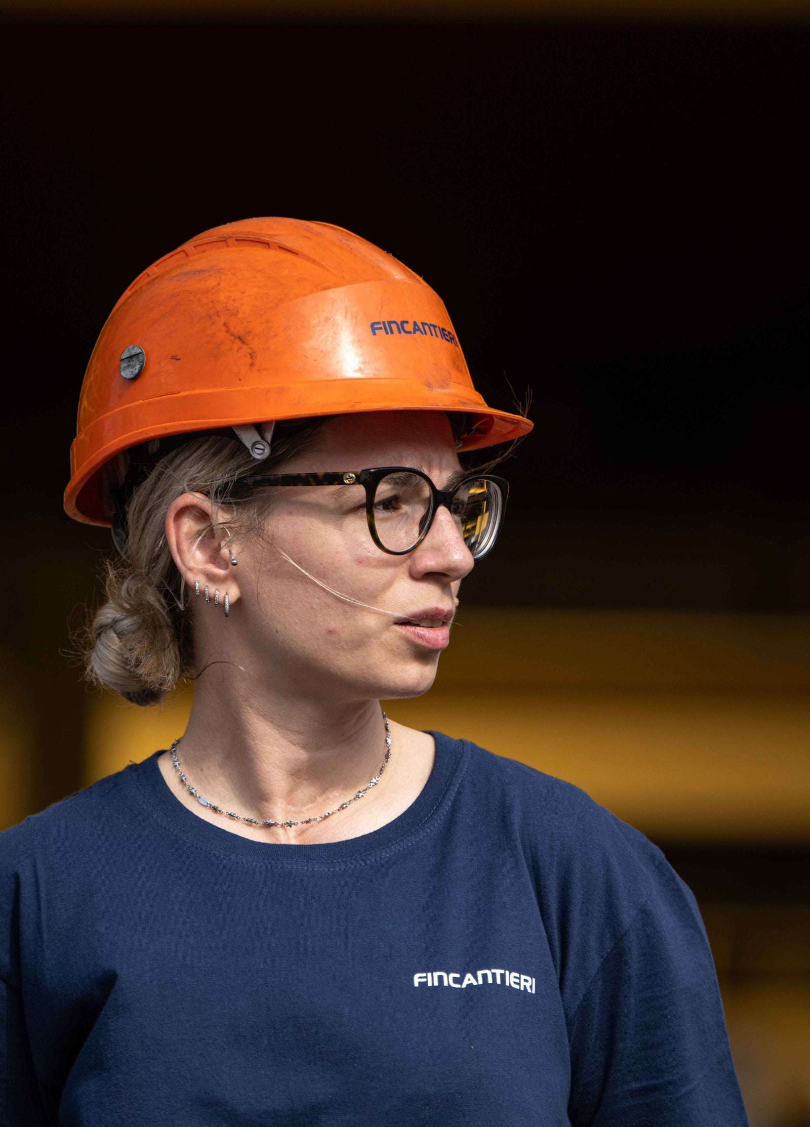 Fincantieri | Group - Sustainability - Industrial Excellence - Health and Safety: Person wearing an orange Fincantieri helmet and blue shirt with visible Fincantieri text, industrial or shipyard environment with warm lighting, personal details such as necklace and glasses.