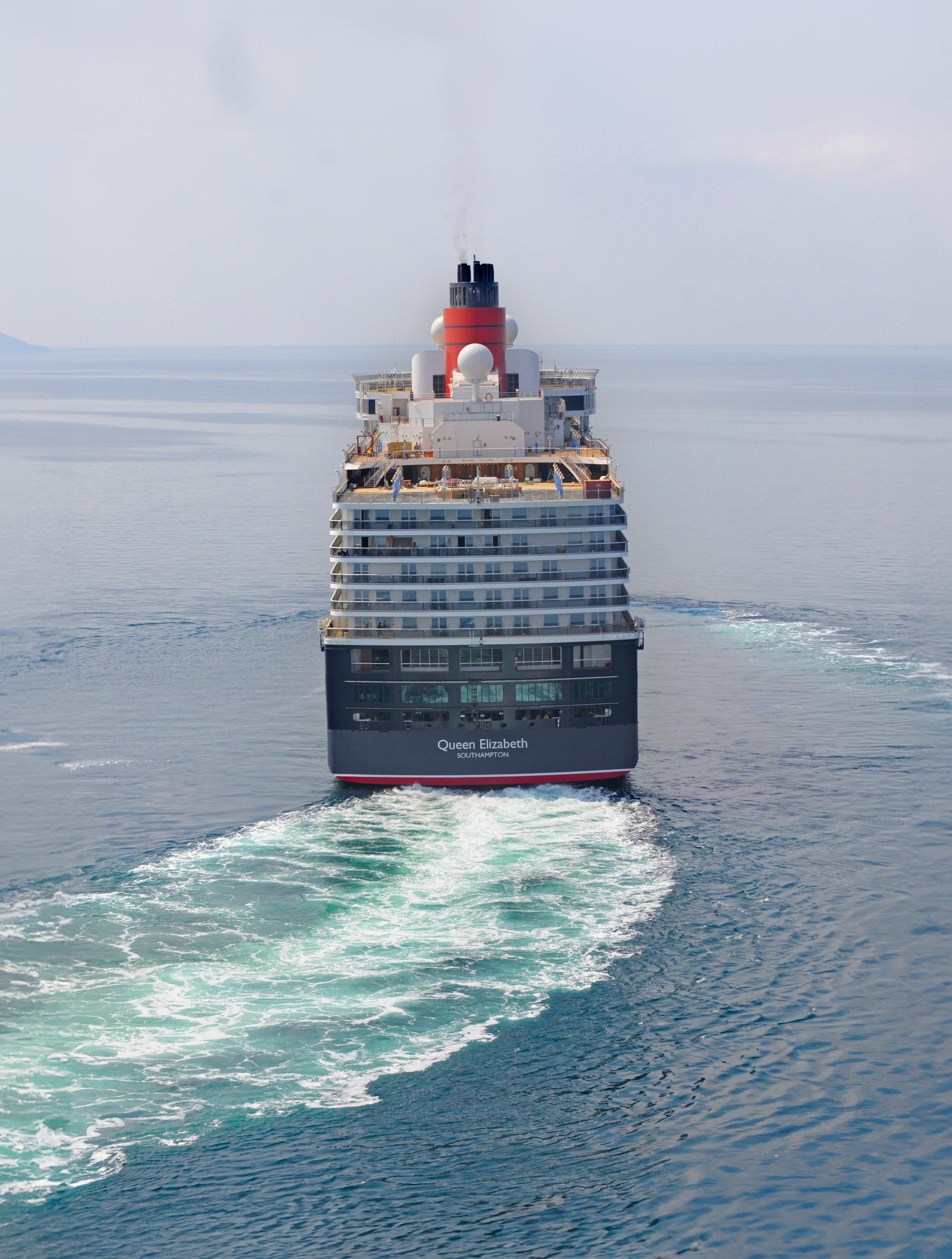 Fincantieri | Group - Business - Products - Cruise Ships - Queen Elizabeth: Rear view of Queen Elizabeth cruise ship in operation navigating open sea, visible white wake, 'Queen Elizabeth' name on stern hull, red and black funnel, distant land barely visible on horizon.