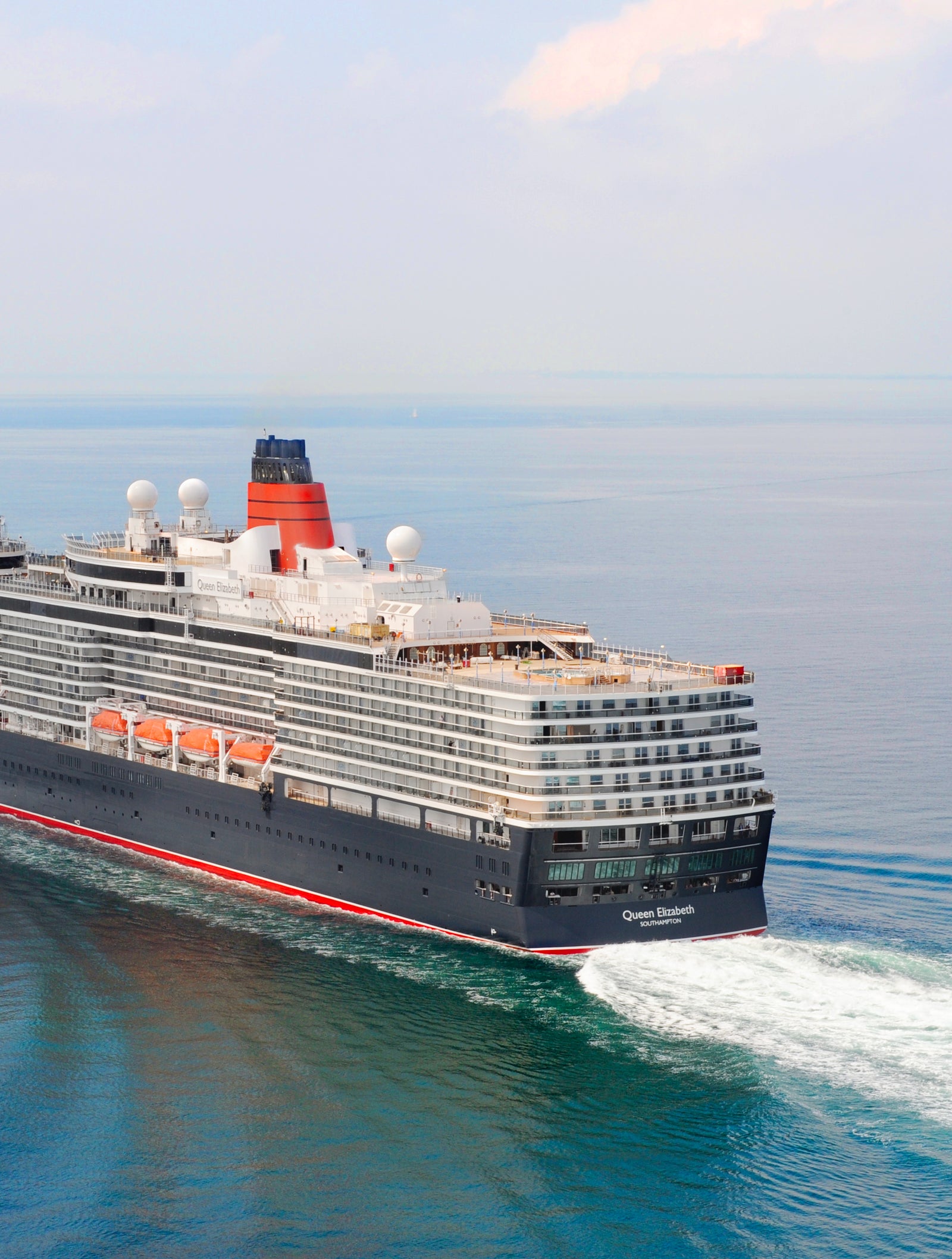 Fincantieri | Group - Business - Products - Cruise Ships - Queen Elizabeth: Rear side view of Queen Elizabeth cruise ship in operation navigating open sea, multiple passenger decks, visible lifeboats along sides, hull with red and black design.